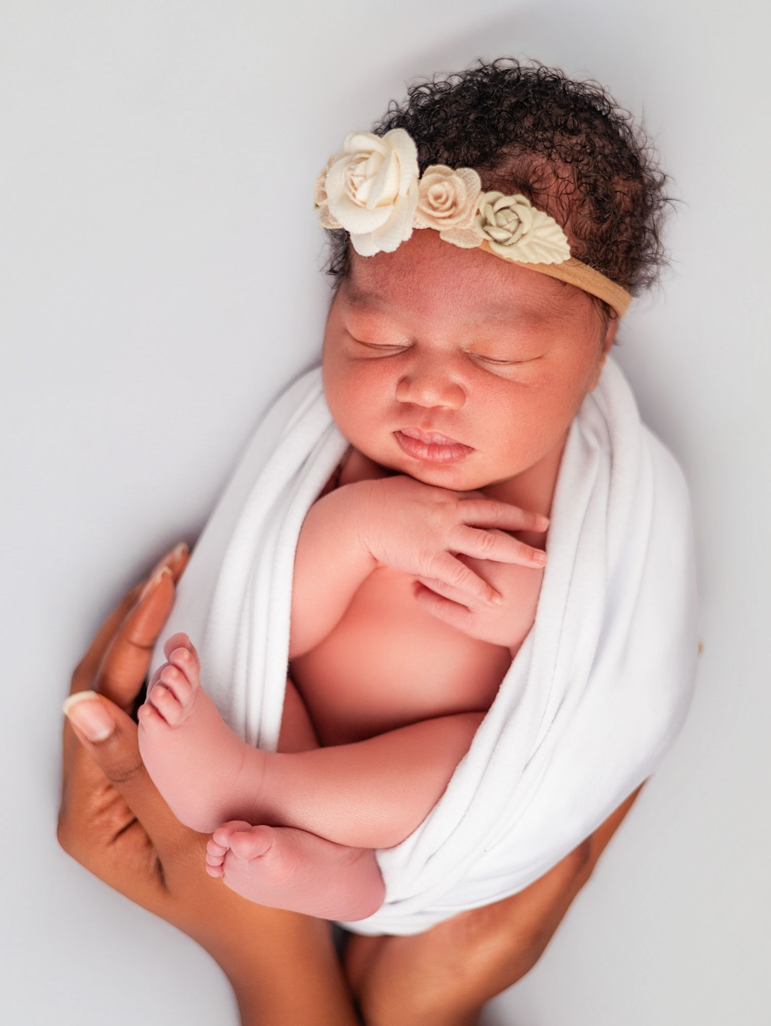 Newborn baby girl wrapped in white swaddle with floral headband, cradled in hands against gray background.