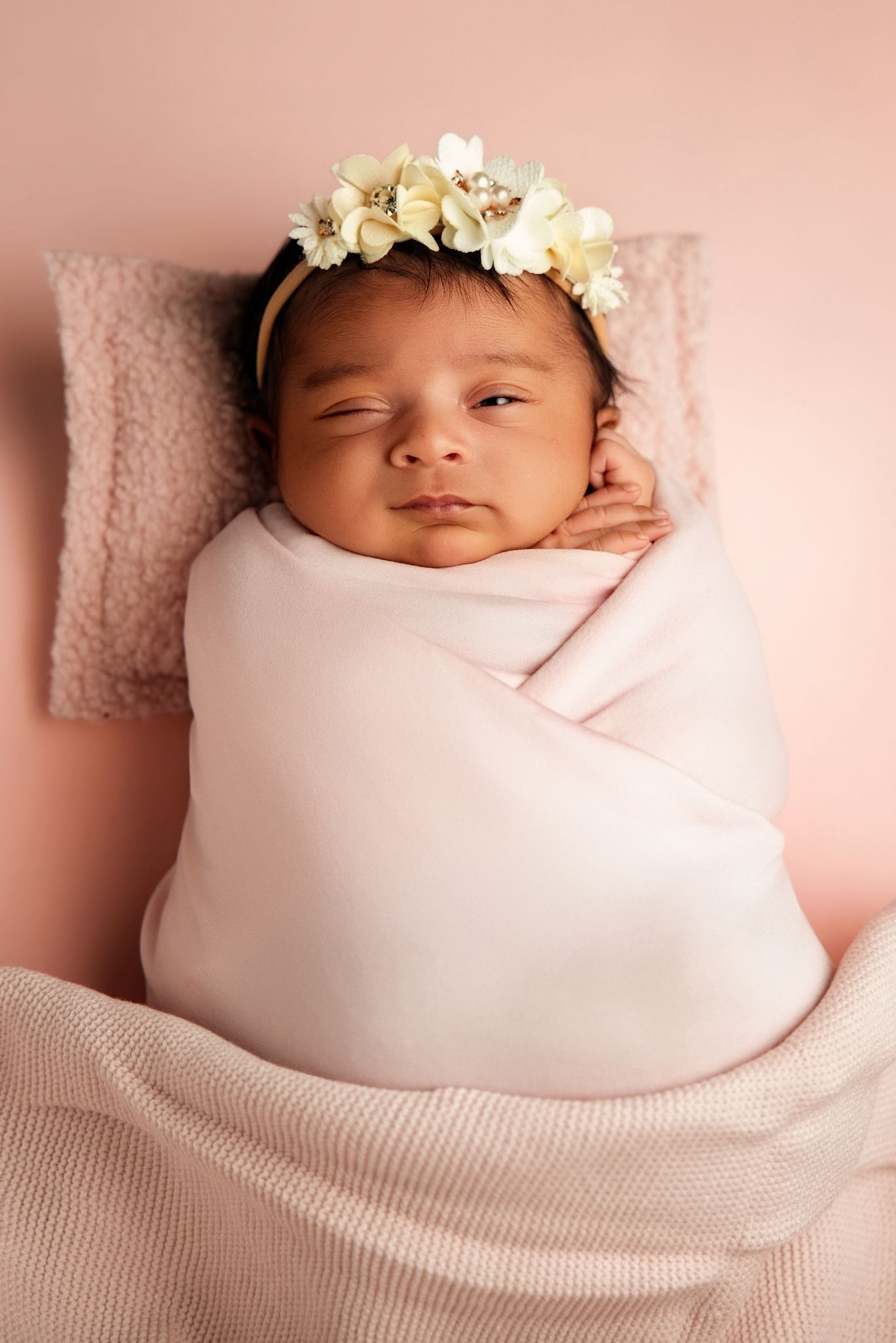 Newborn baby girl swaddled in pink blanket wearing floral crown, peacefully resting on pink pillow.
