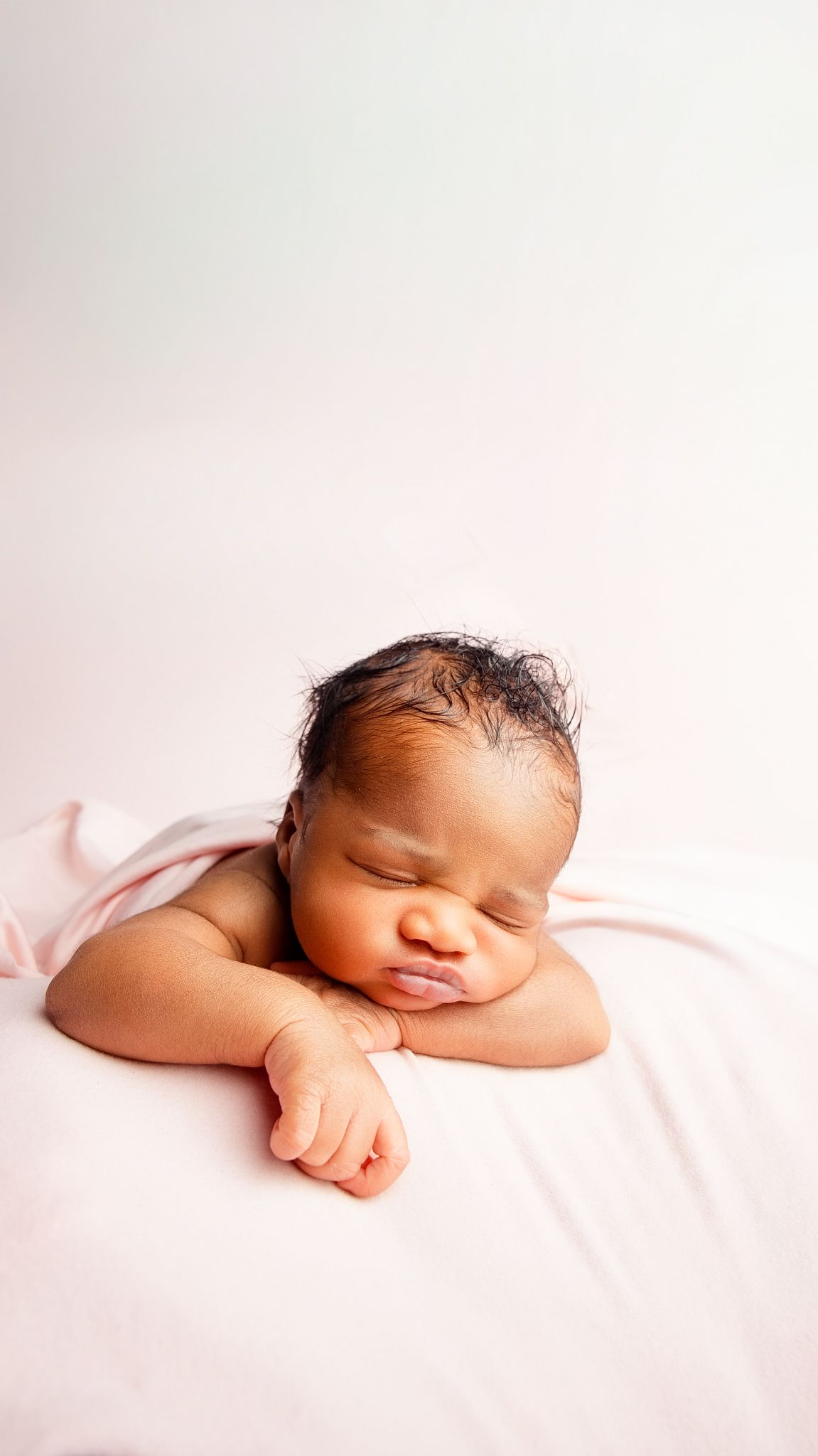 Peaceful newborn baby sleeping on a soft pink blanket with hands folded under chin against a white background.