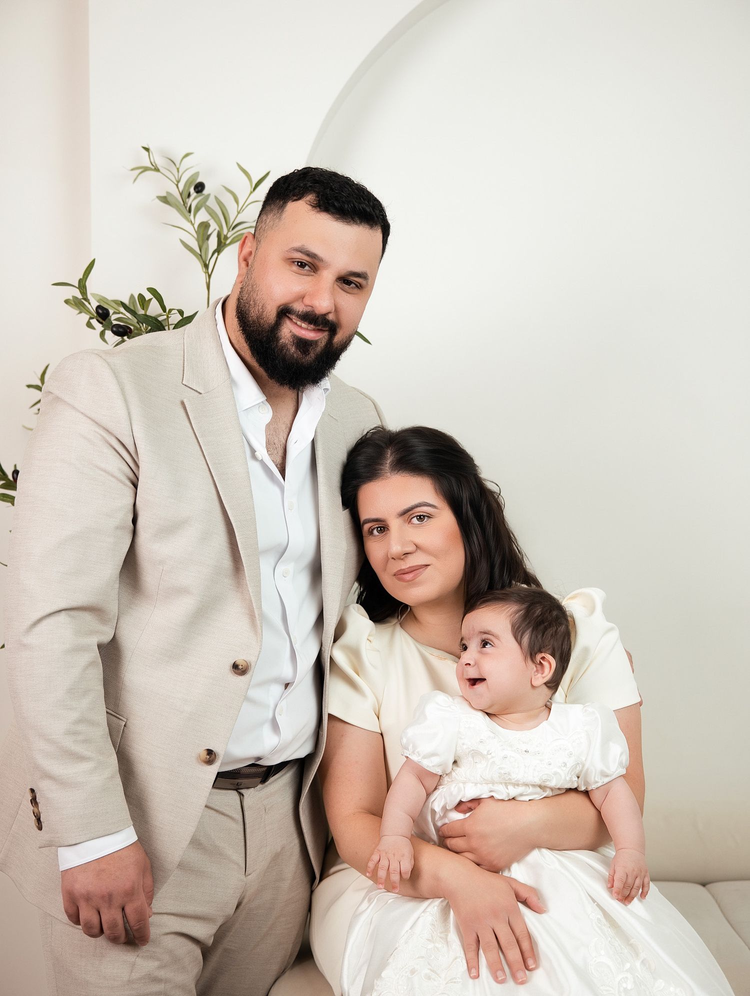 Smiling couple in formal attire poses with their infant daughter in a white lace outfit in Family portrait photographer Downtown Toronto