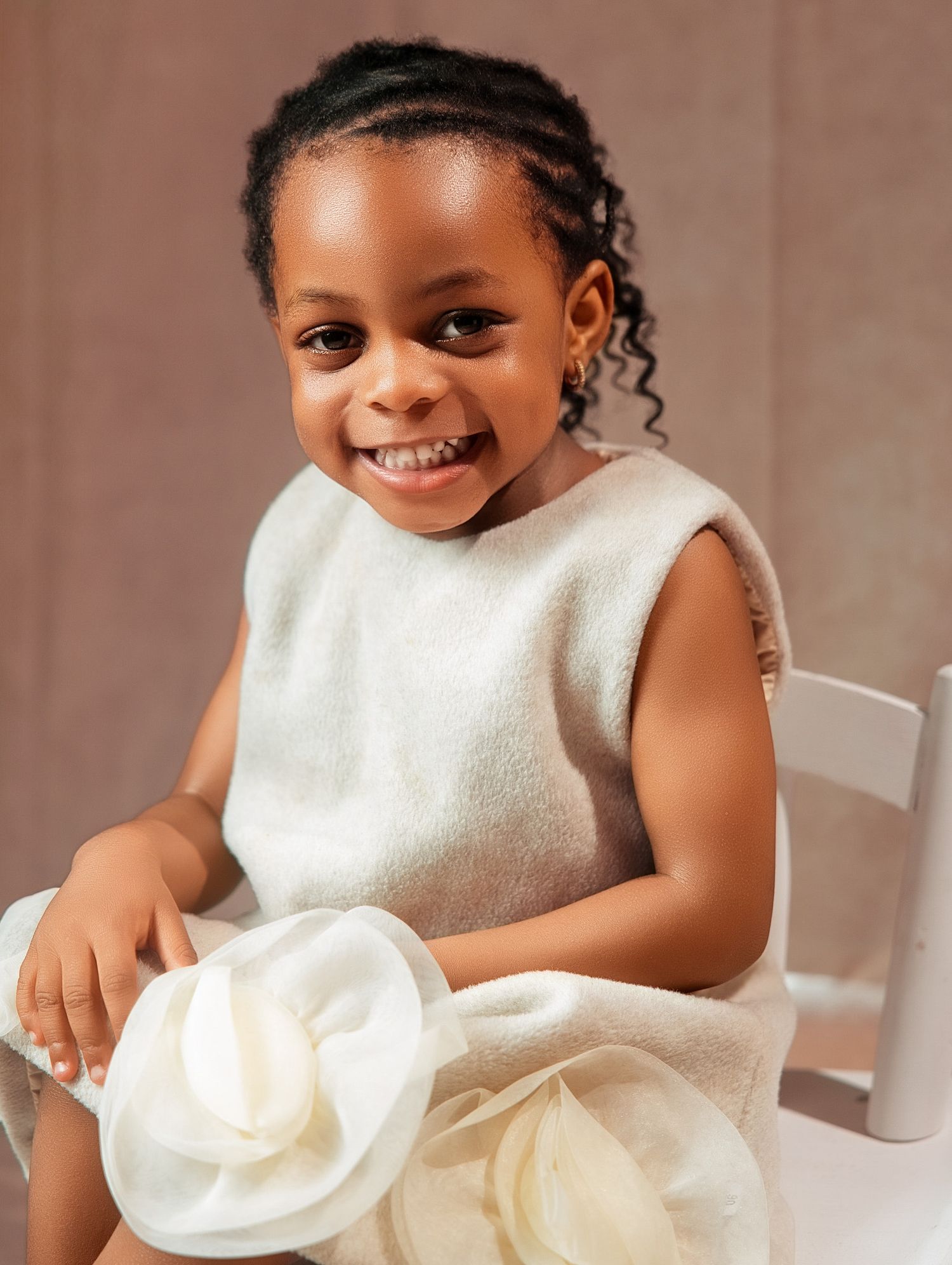 Smiling young Black girl in white sleeveless dress holding accessory, seated on white chair captured by a baby's photographer in North York
