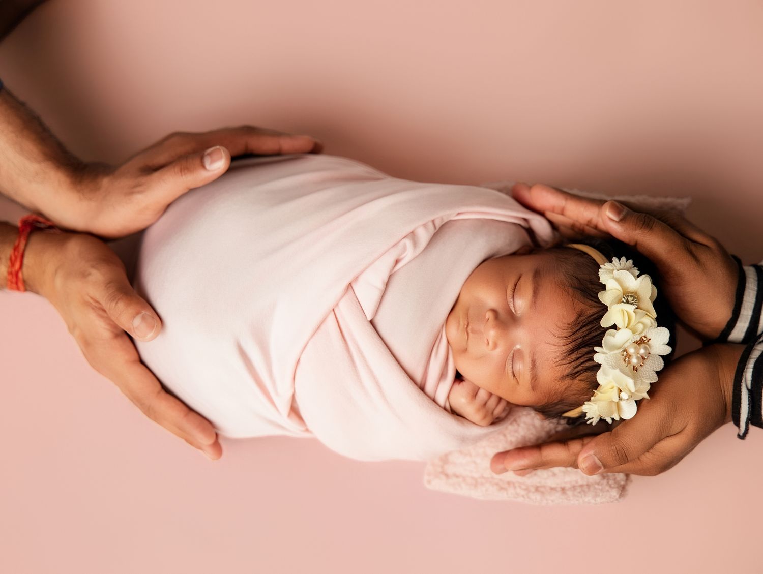 Newborn baby girl wrapped in pink swaddle with floral headband, cradled by parents hands on pink background Newborn photography in Toronto