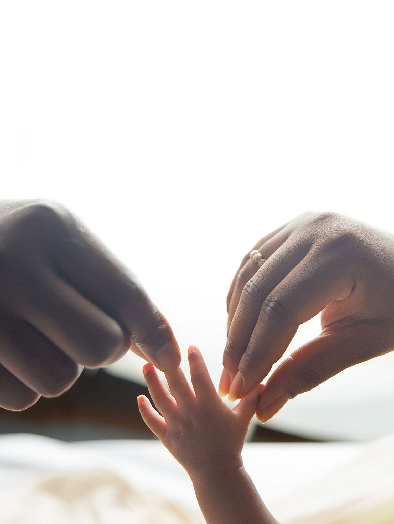 Adult hands gently touching a newborn baby's tiny fingers in a tender, backlit moment of connection.