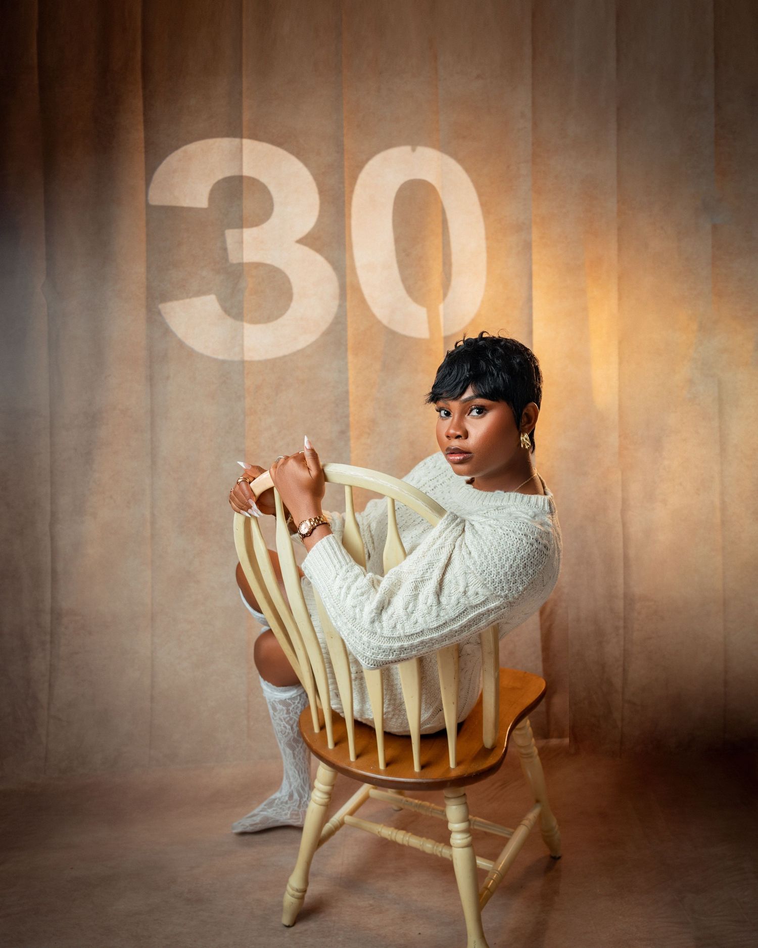 Young woman in cream cable-knit sweater sits on wooden chair before illuminated number 30 backdrop in toronto birthday photography
