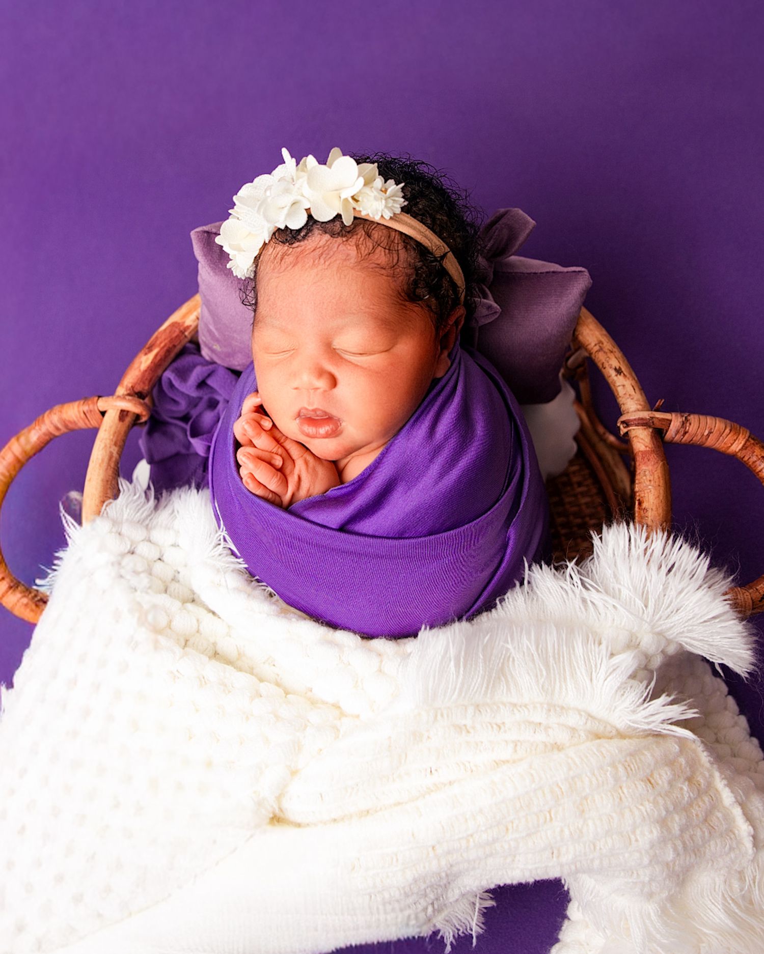 Sleeping newborn wrapped in purple swaddle with white flower headband in wicker basket Newborn photography in Toronto