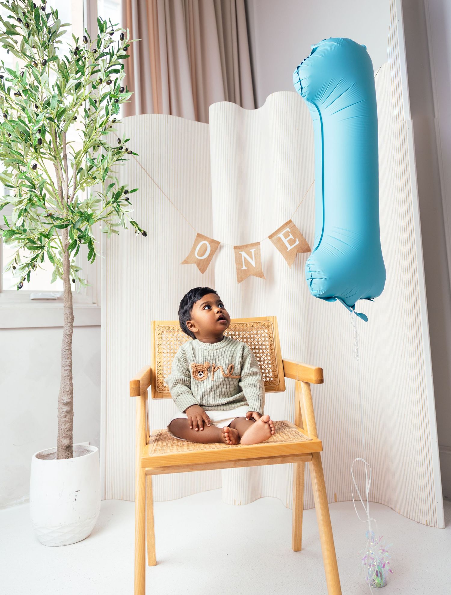 A toddler boy sits on a wooden chair looking up at a blue number one balloon at his first birthday.