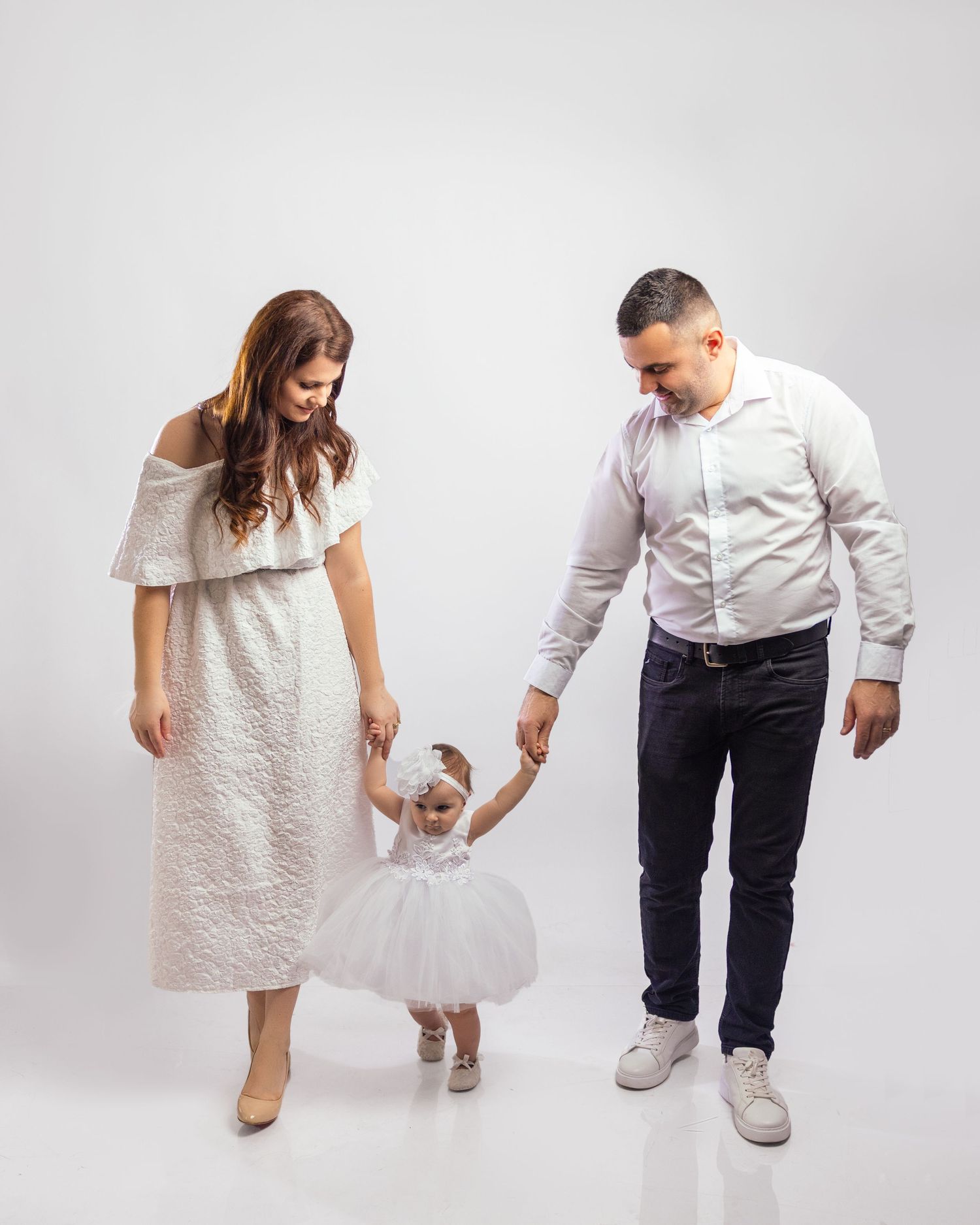 Family portrait of parents in white outfits helping their toddler daughter walk in a white toronto family studio setting.