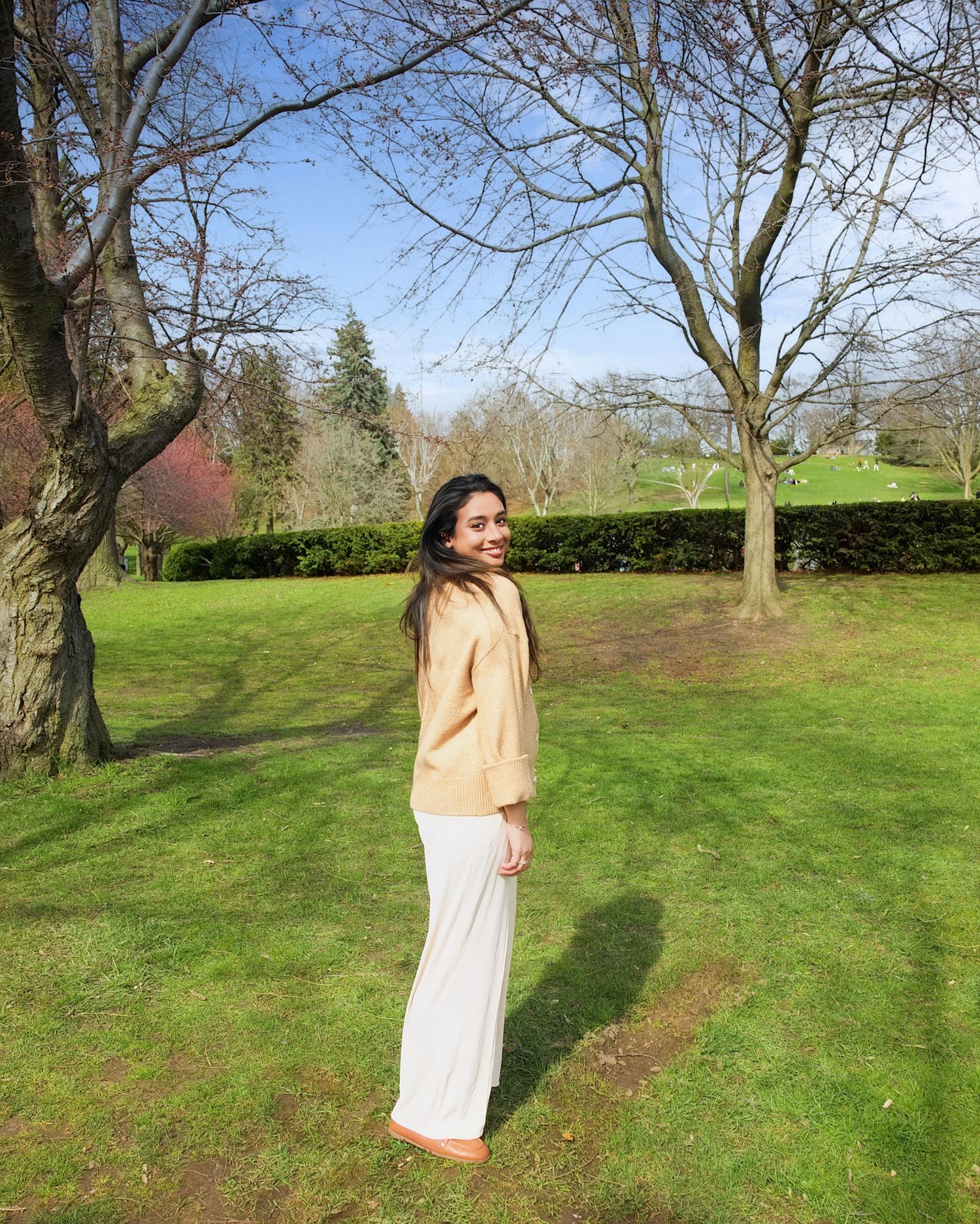 Young woman in yellow top and white wide-leg pants standing in a sunny green park with bare trees in relaxed outdoor birthday photos toronto