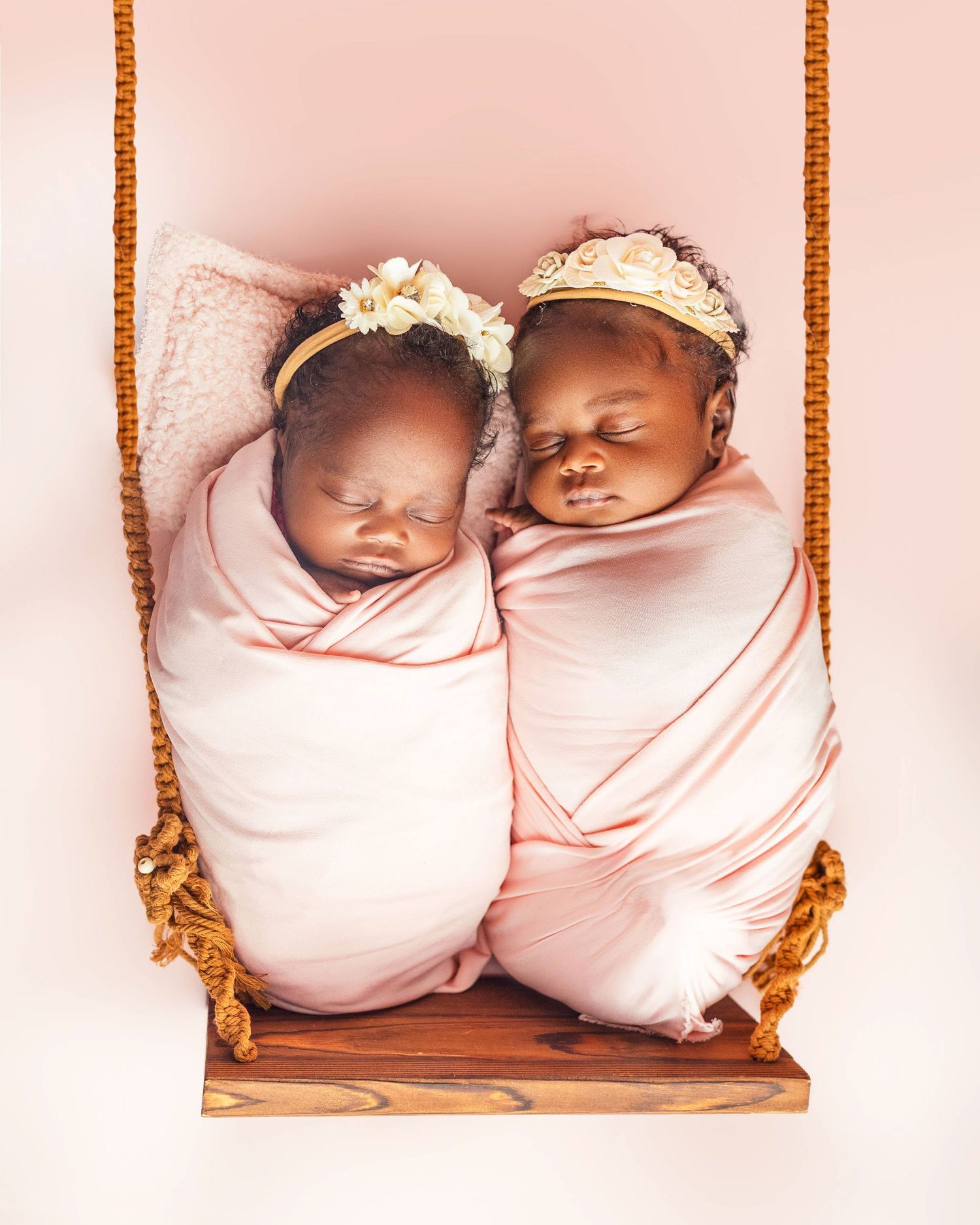Two swaddled newborn twin girls sleeping on a wooden swing, wearing floral headbands against a pink background.