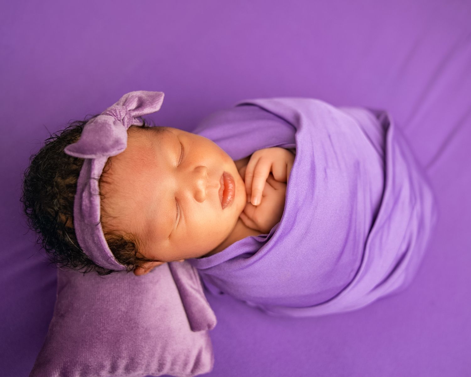 Sleeping newborn baby wrapped in purple swaddle with bow headband on matching purple background.