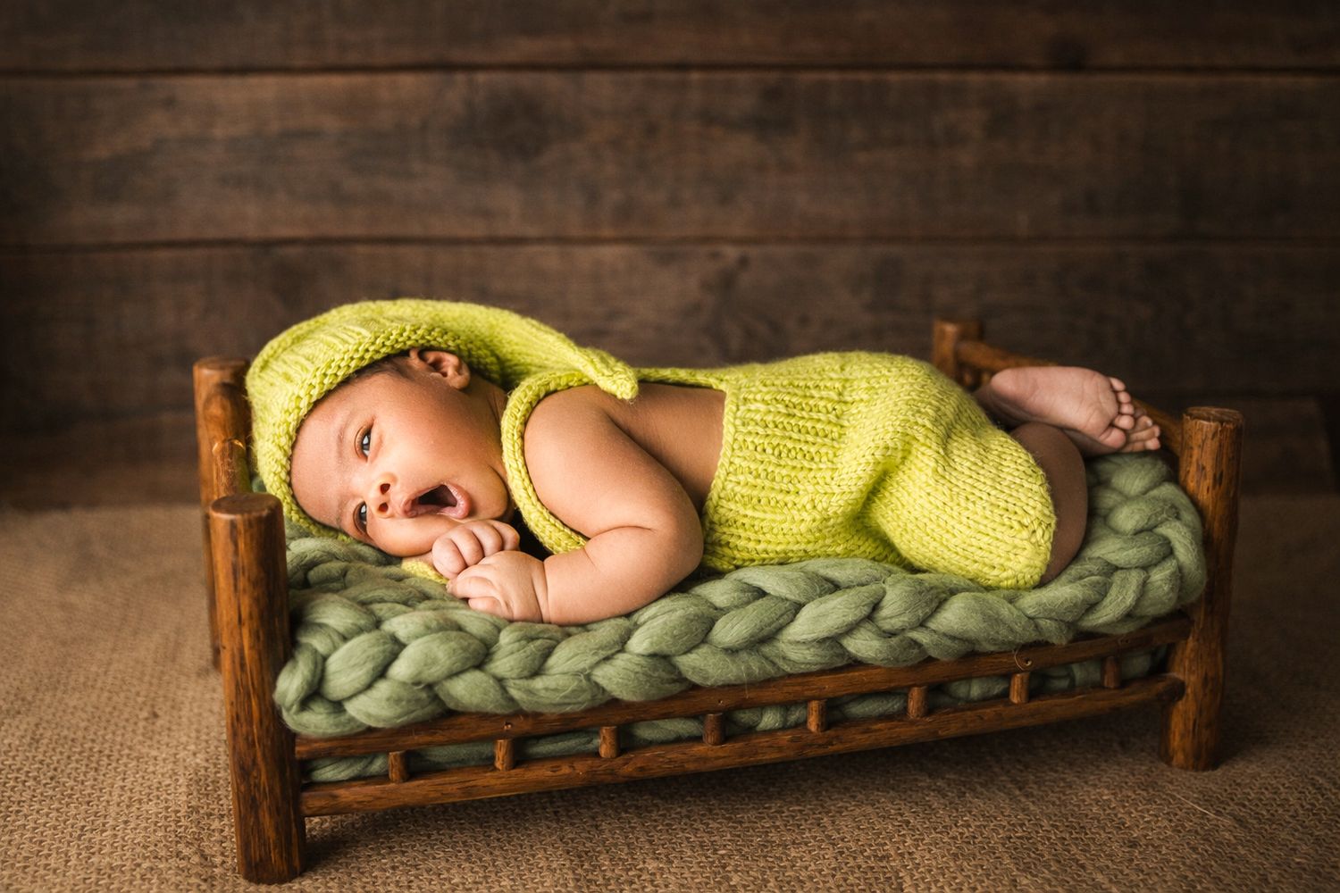 Newborn baby in green knit outfit lying on a miniature wooden bed with green braided blanket captured by Family photography near Downtown To