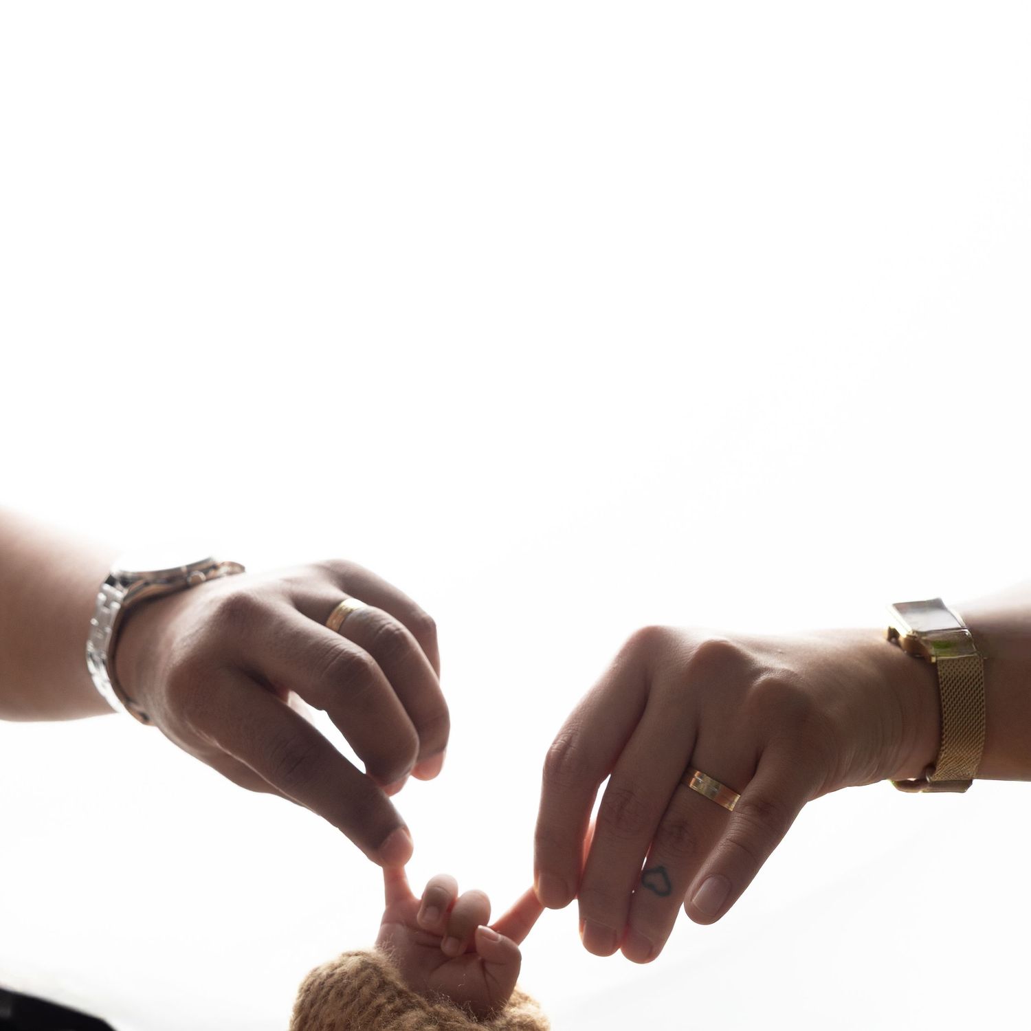 Two adults wearing rings and watches gently hold a newborn baby's tiny feet with their fingertips.