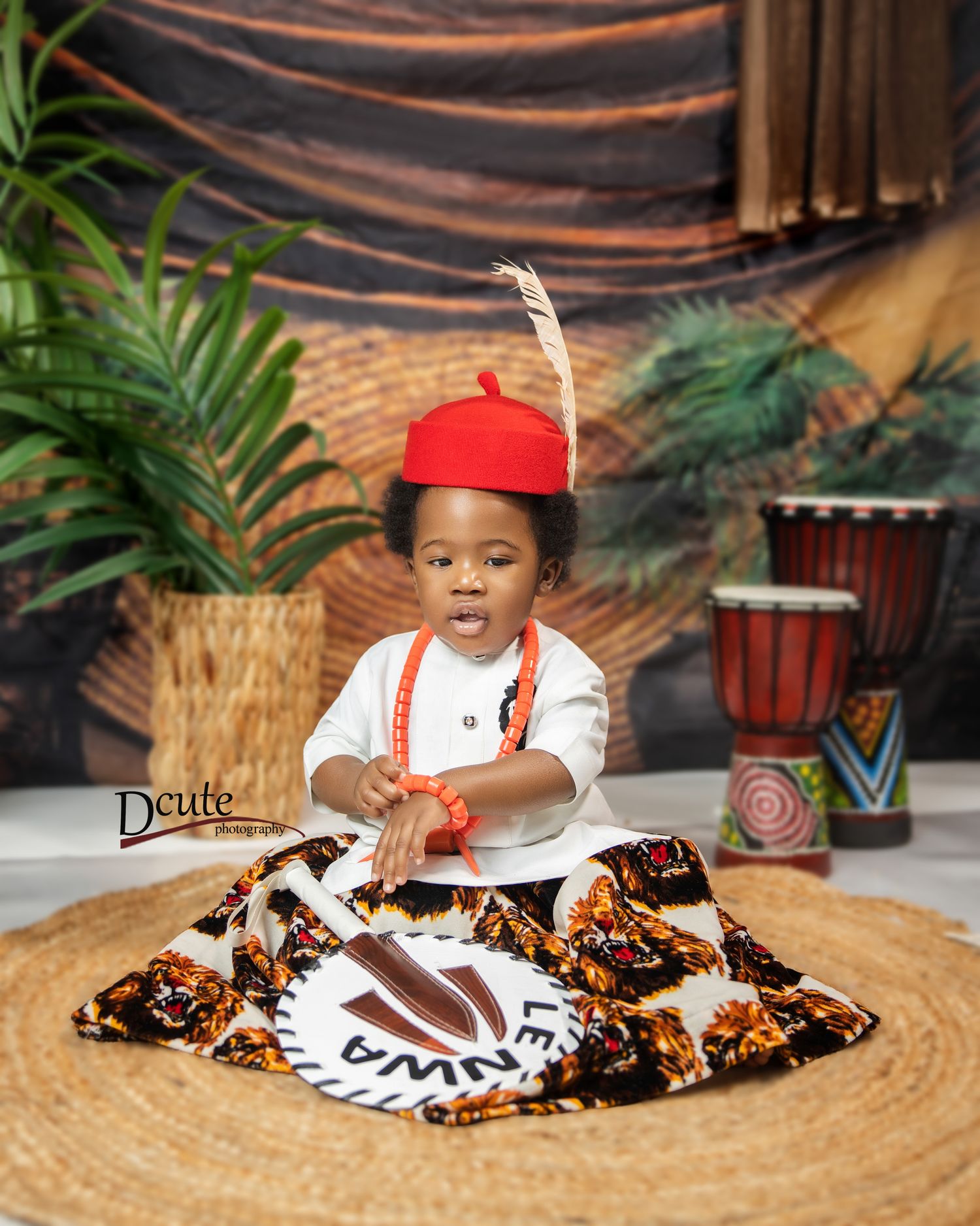 Adorable toddler in traditional African attire with red hat and feather sitting on a woven mat with a Children’s photographer in North York