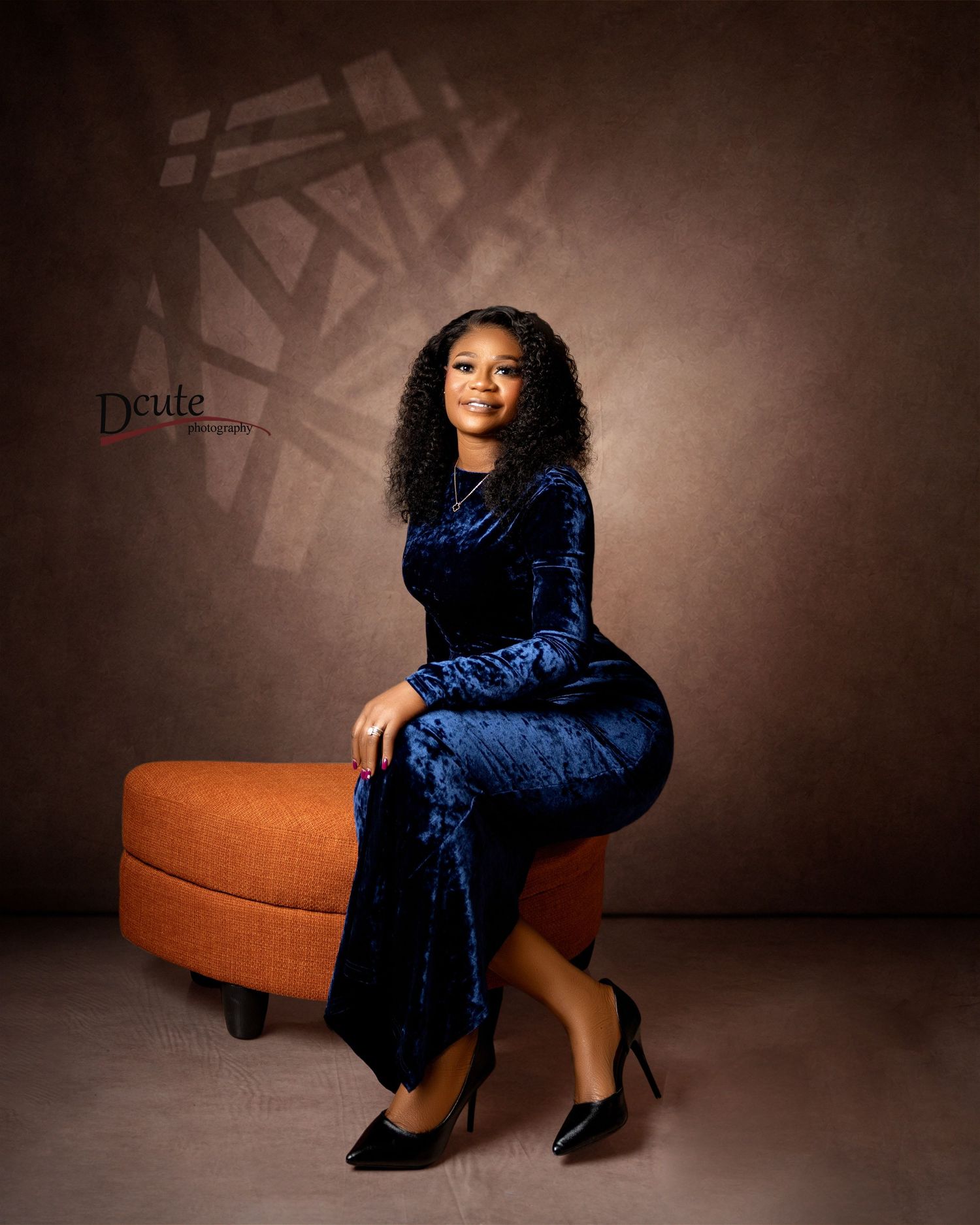 Woman in blue velvet dress seated on orange ottoman in professional studio birthday portrait photoshoot
