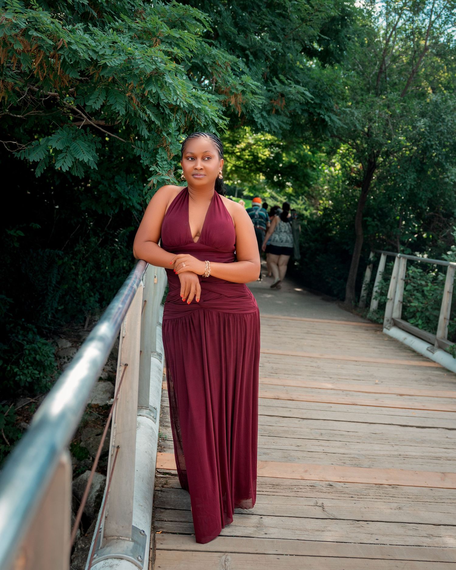 Woman in elegant burgundy halter maxi dress poses on wooden bridge surrounded by lush green trees in toronto outdoor birthday photography