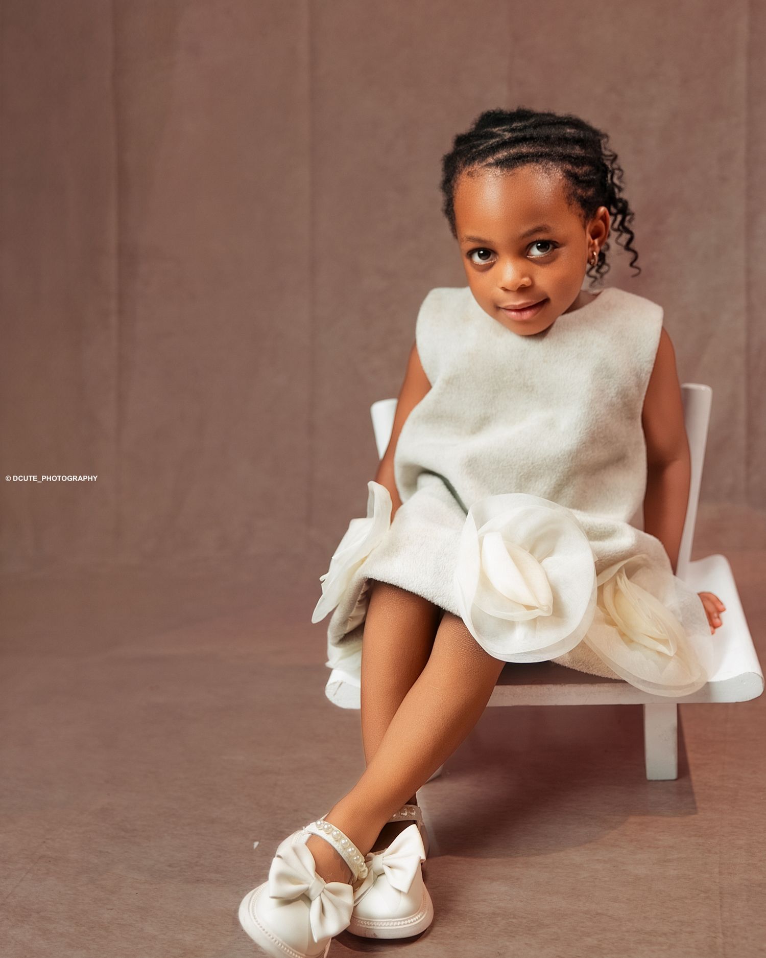 Young Black girl in white dress with floral detail seated on small white chair wearing bow taken by north york kids photographer