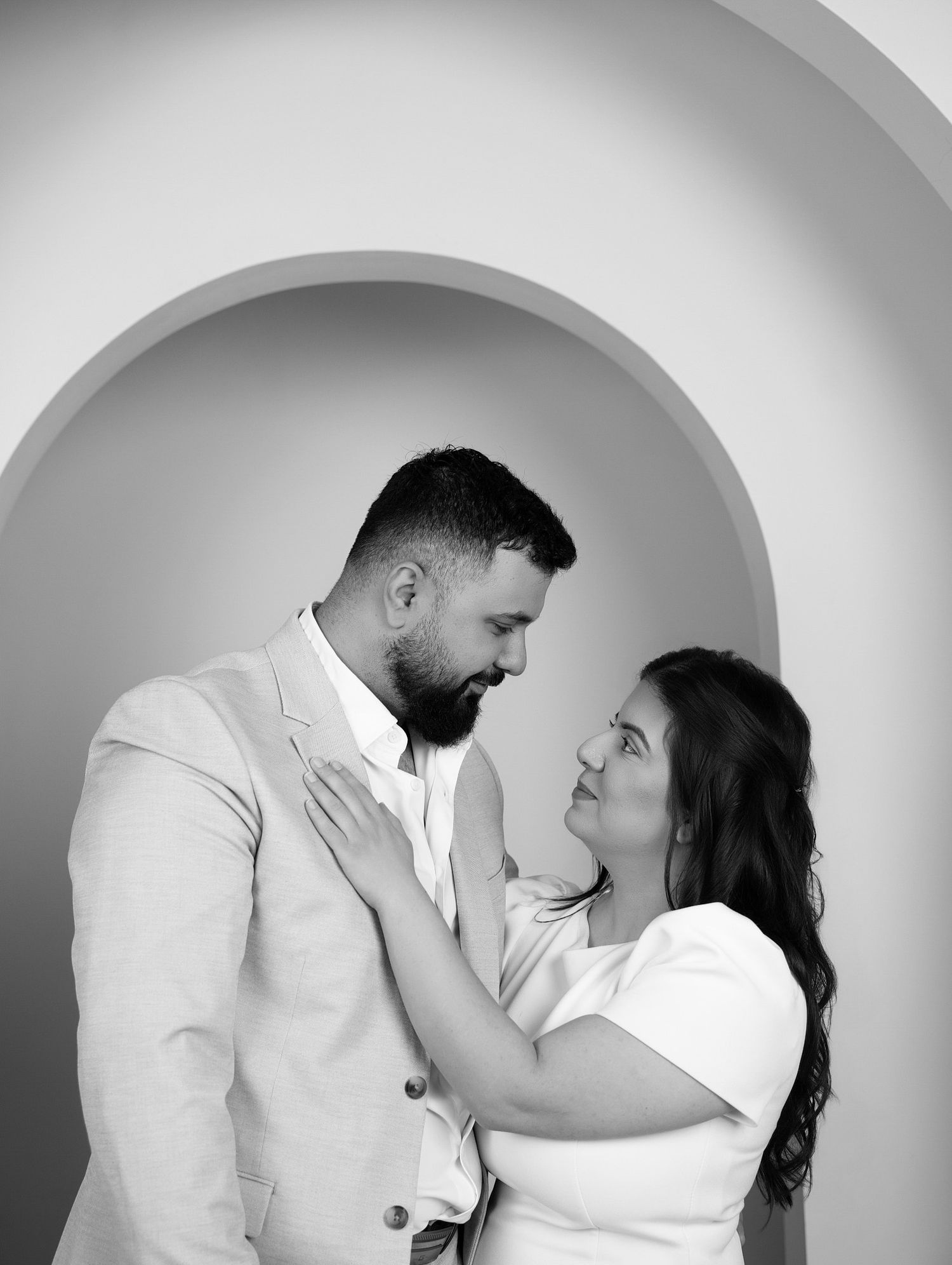 A couple in black and white photo gazes at each other tenderly under an elegant arched doorway.