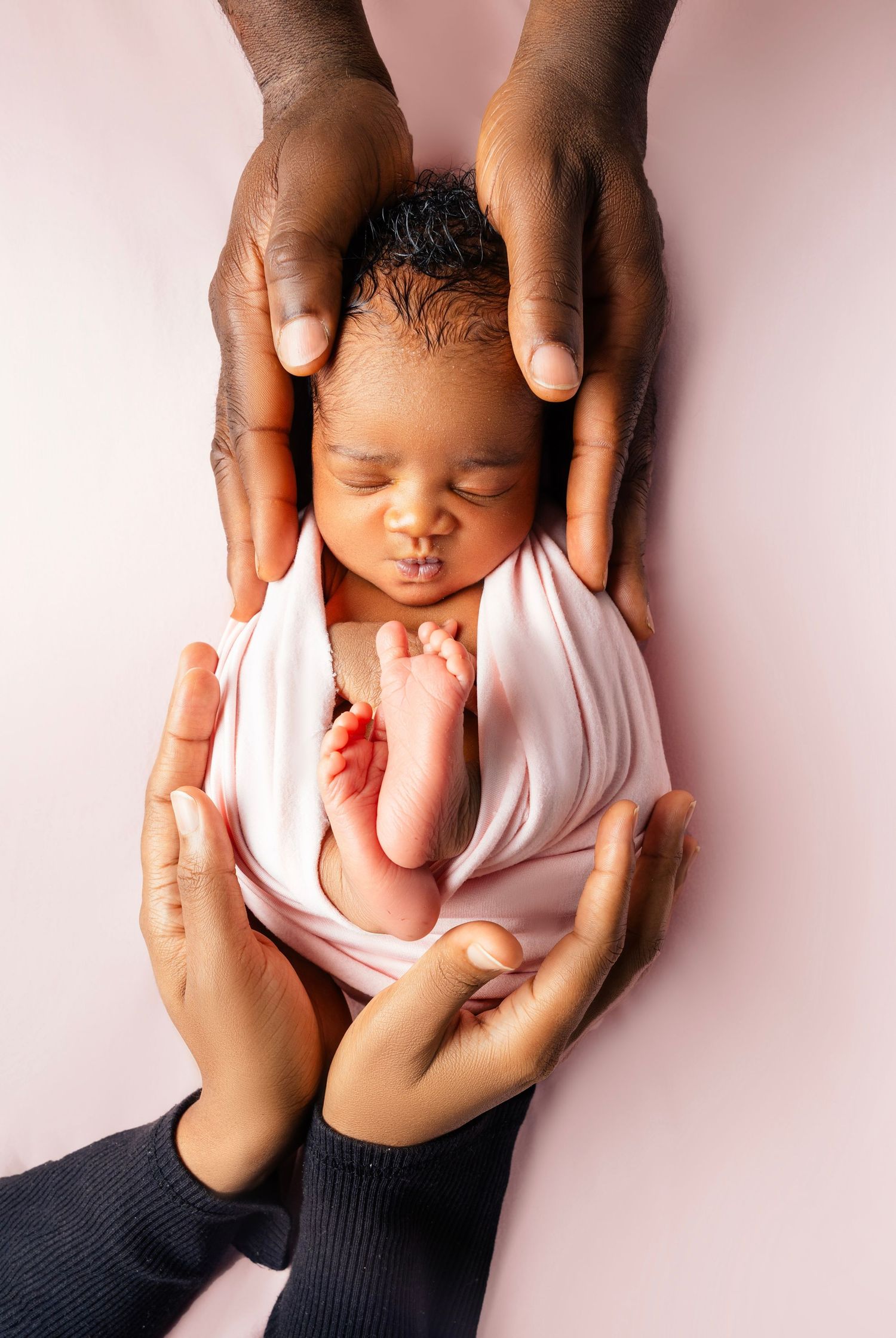 Newborn baby swaddled in pink wrap, cradled gently by two pairs of parents hands on a soft pink background.