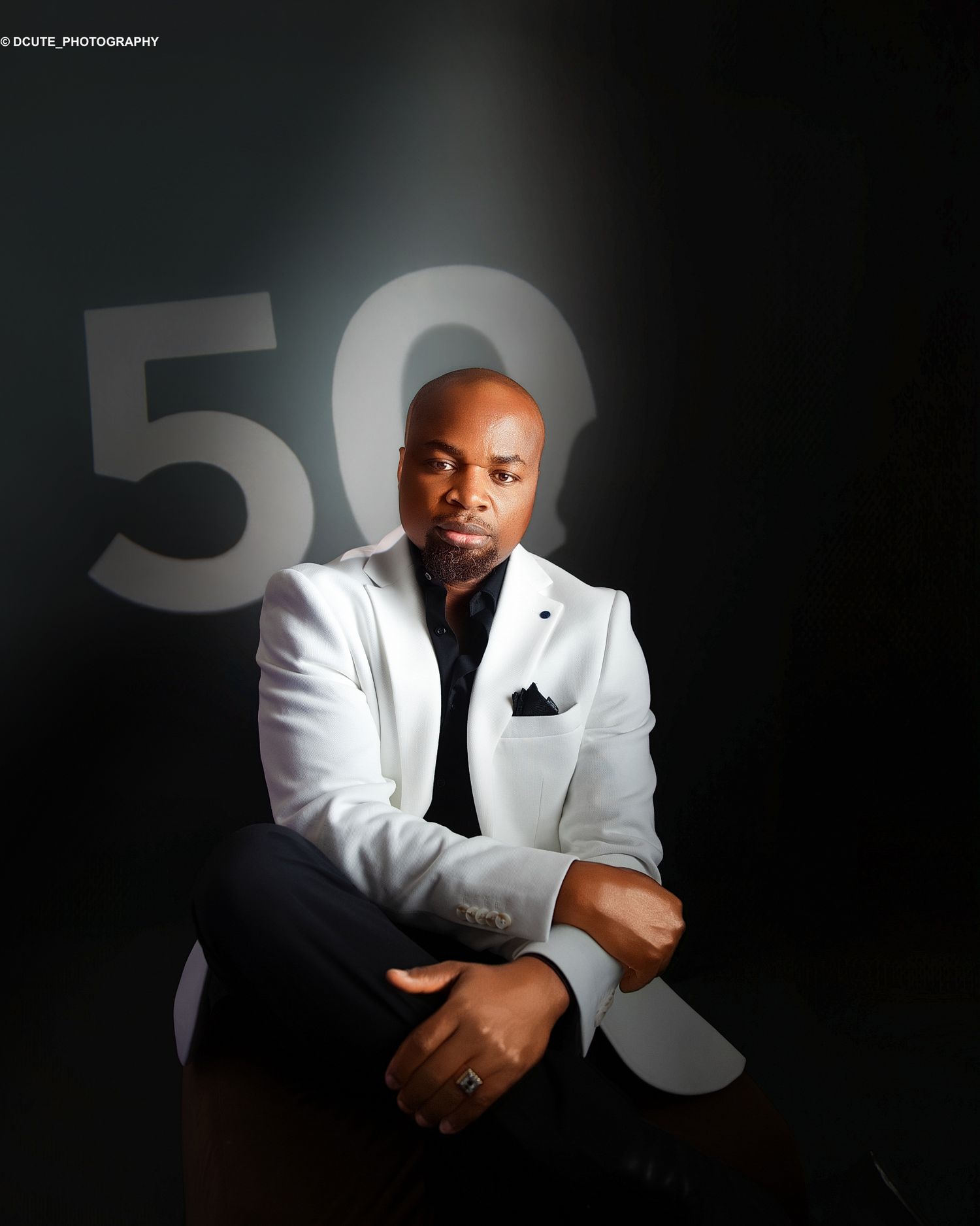 Man in white blazer and black outfit seated against dark background with large number 50 during birthday photoshoot seeion in toronto
