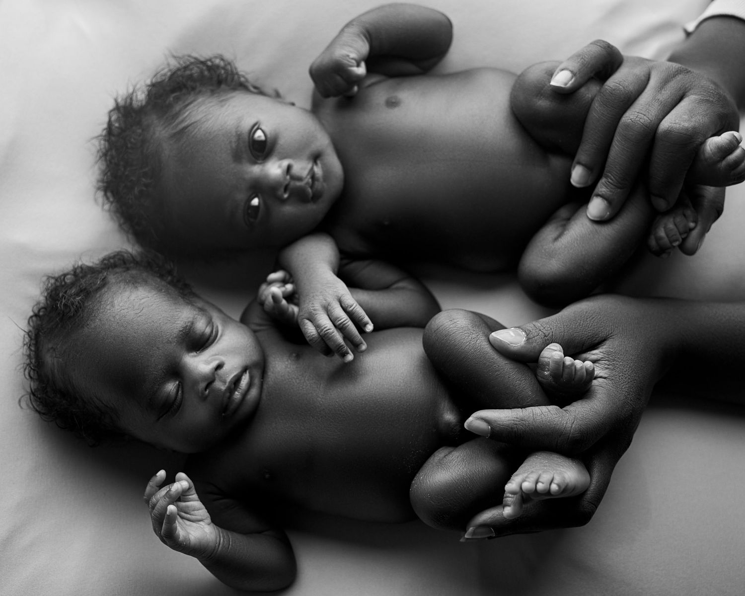 Black and white photo of twin newborn babies in toronto photoshoot lying side by side with an adult hand gently supporting them.