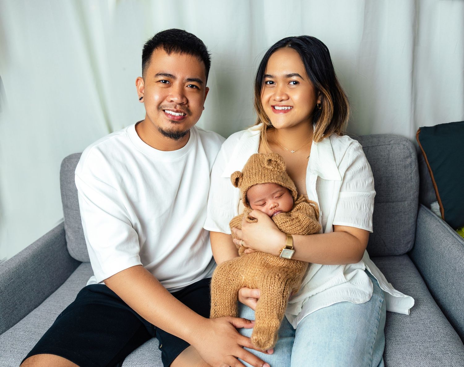 Smiling couple sitting on gray sofa holding their newborn baby dressed in brown bear knit hat in a Newborn photography in Toronto at home