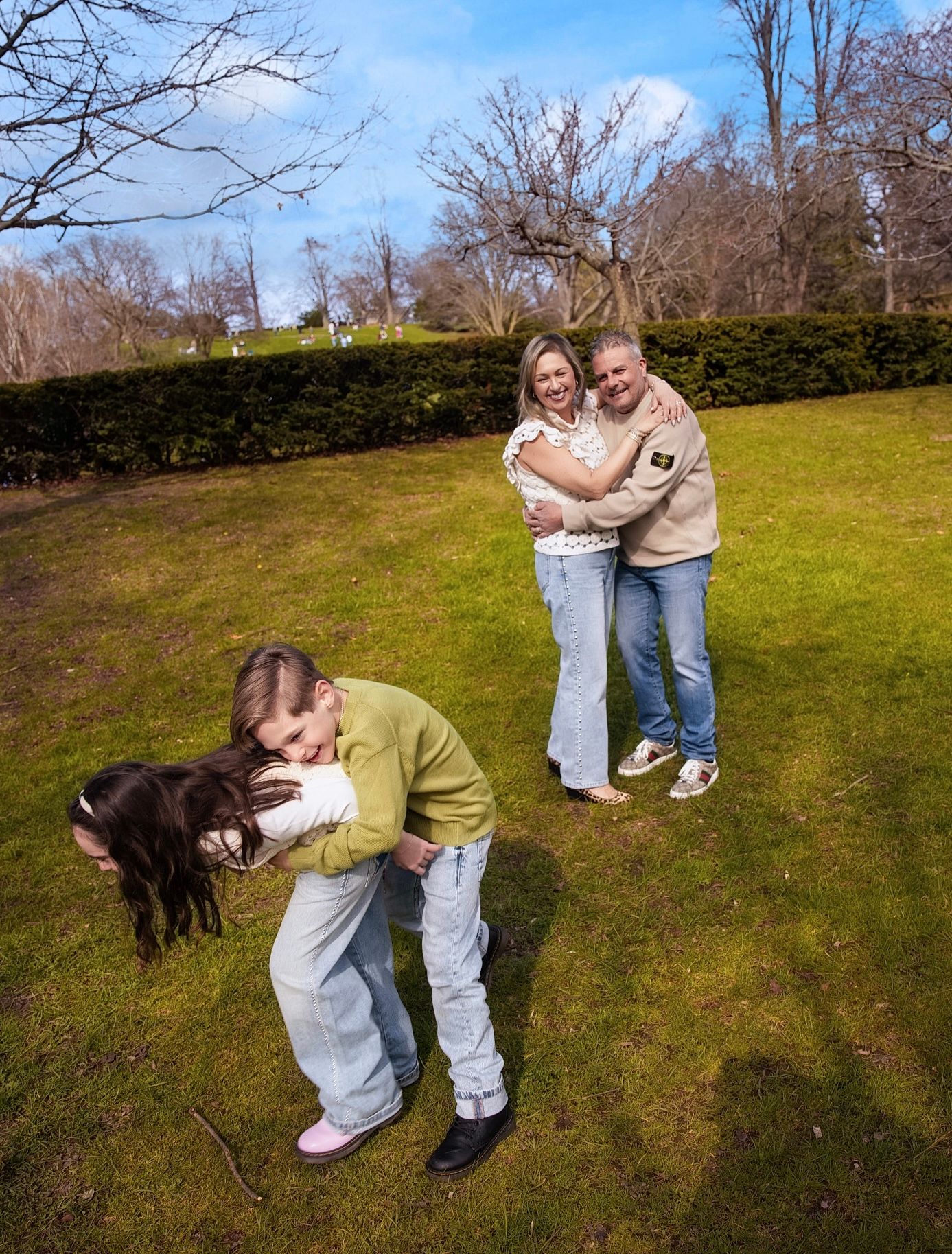 Couple embracing in a sunny park while their daughter plays in the foreground during a candid family photo session in Downtown Toronto