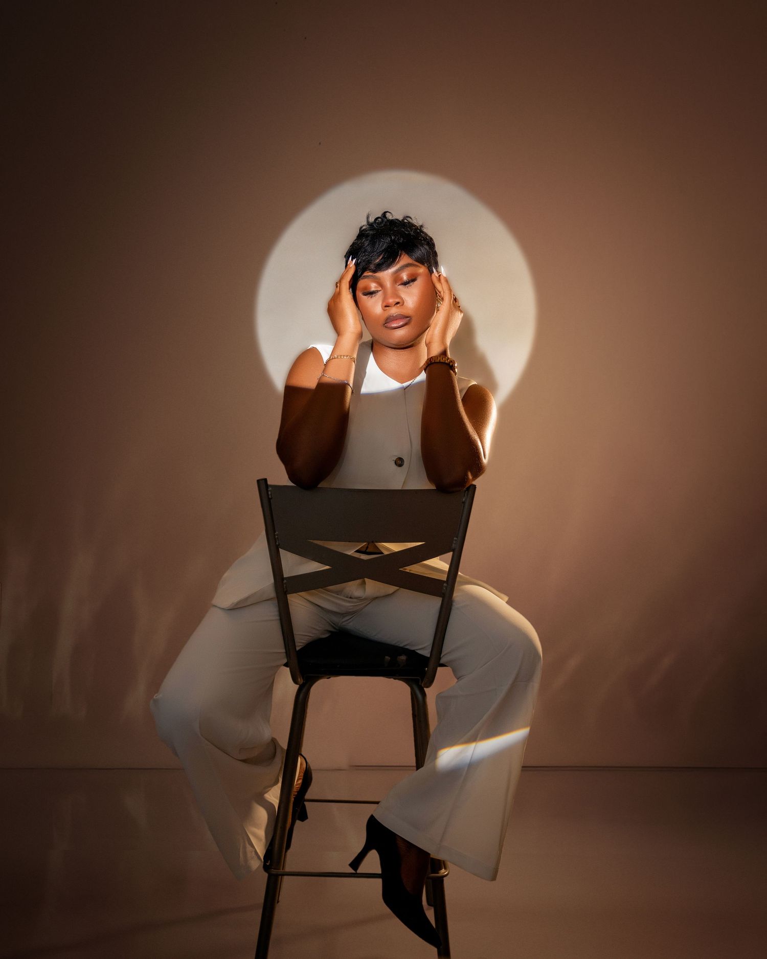 Woman in white and brown outfit sits on chair with hands on temples under dramatic spotlight toronto birthday photography
