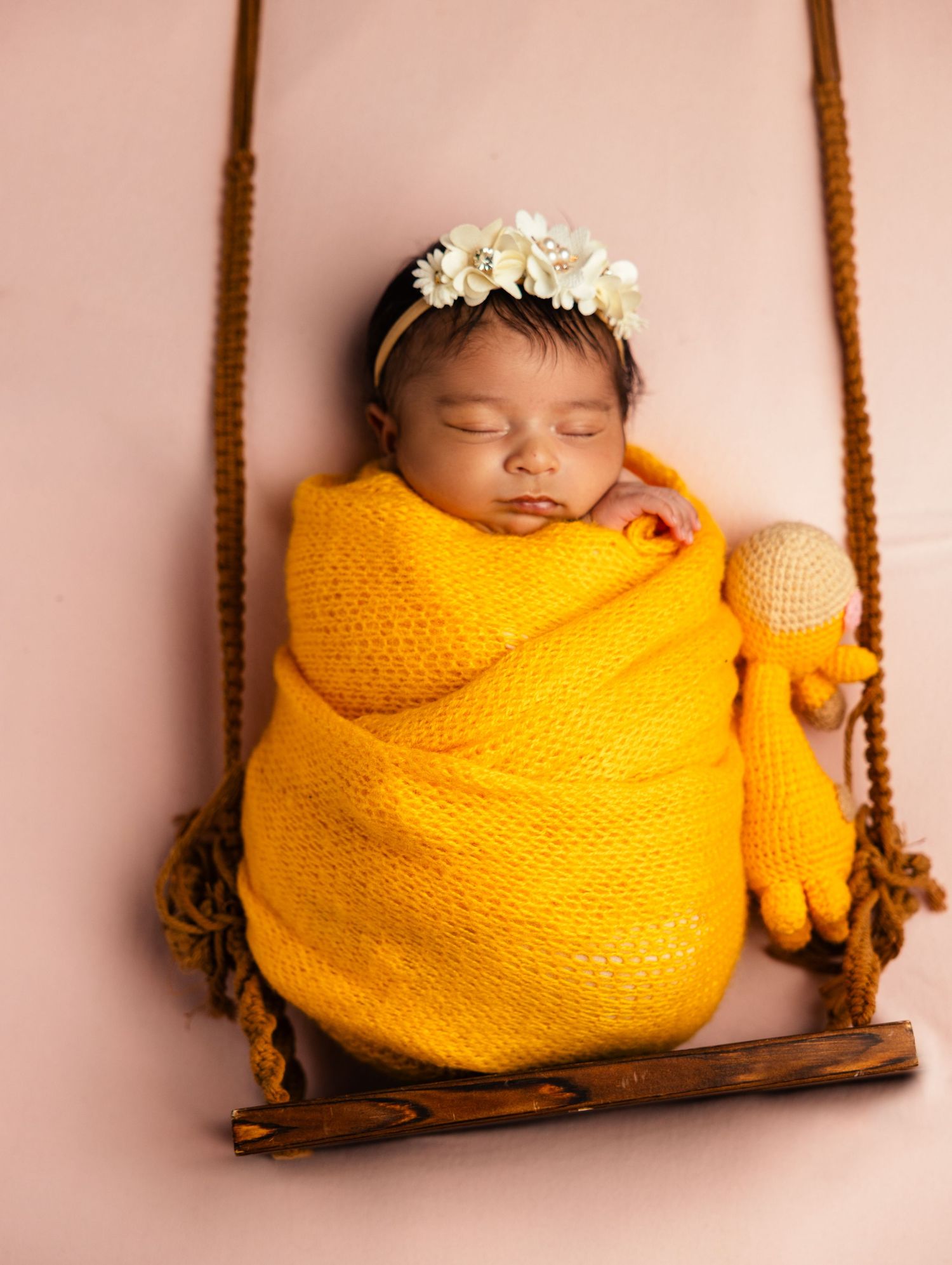A sleeping newborn wrapped in a yellow knit blanket with a floral headband rests on a tiny swing.