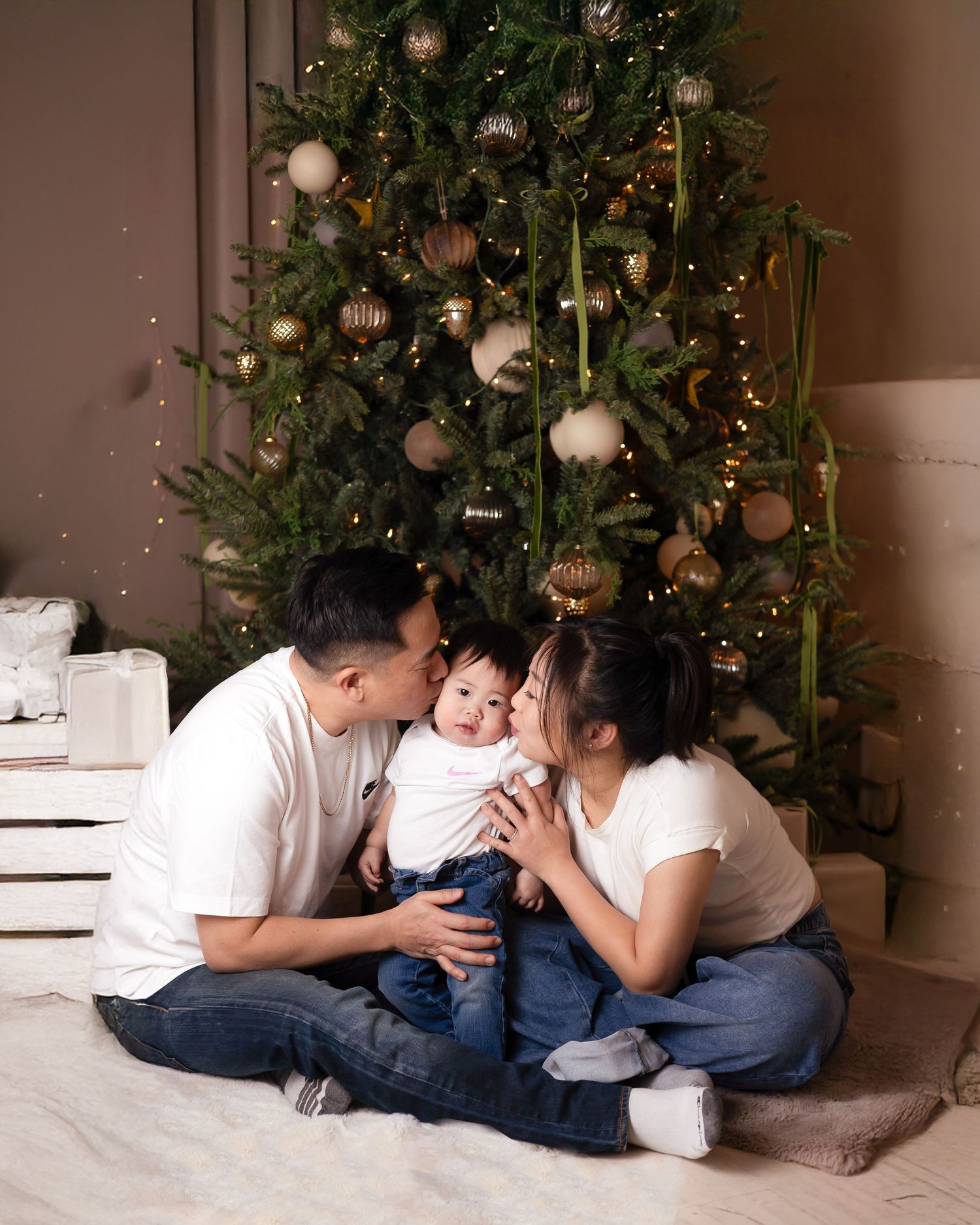 Young Asian family of three share kisses beside a Christmas tree on a white rug in a holiday familly photoshoot near Downtown Toronto