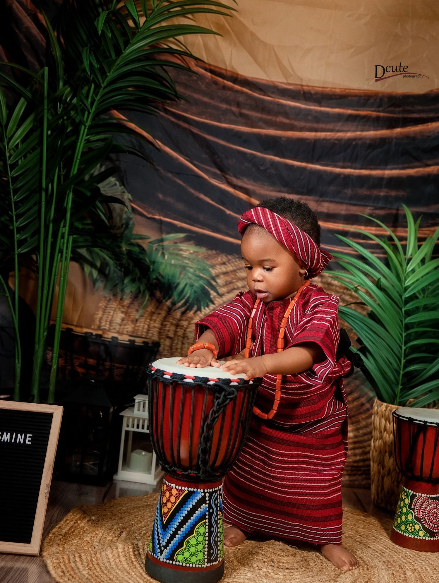 Toddler in red striped traditional outfit plays djembe drum at African-themed first birthday shoot.