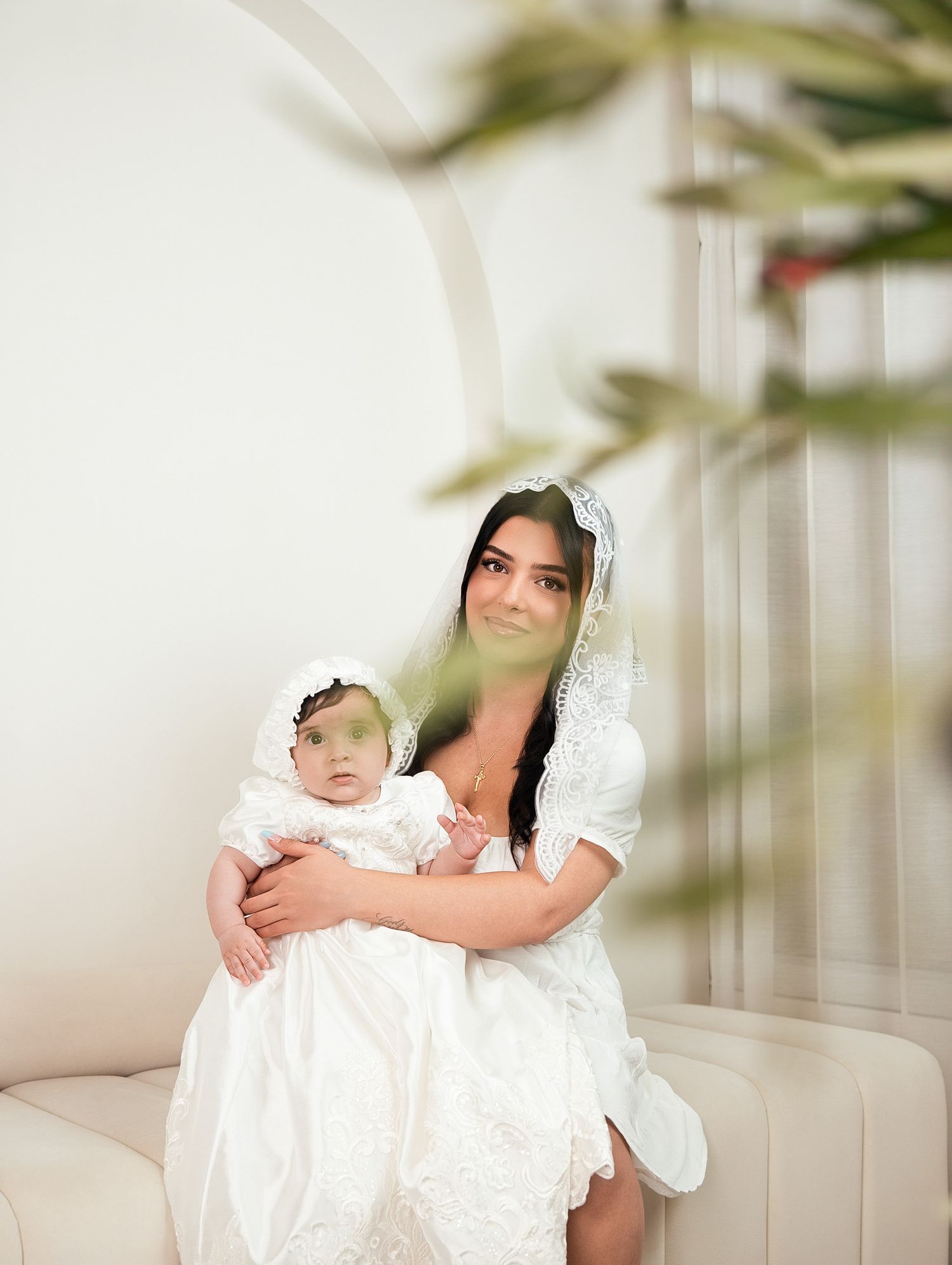 Mother in white dress and lace veil holds baby in christening gown on sofa.