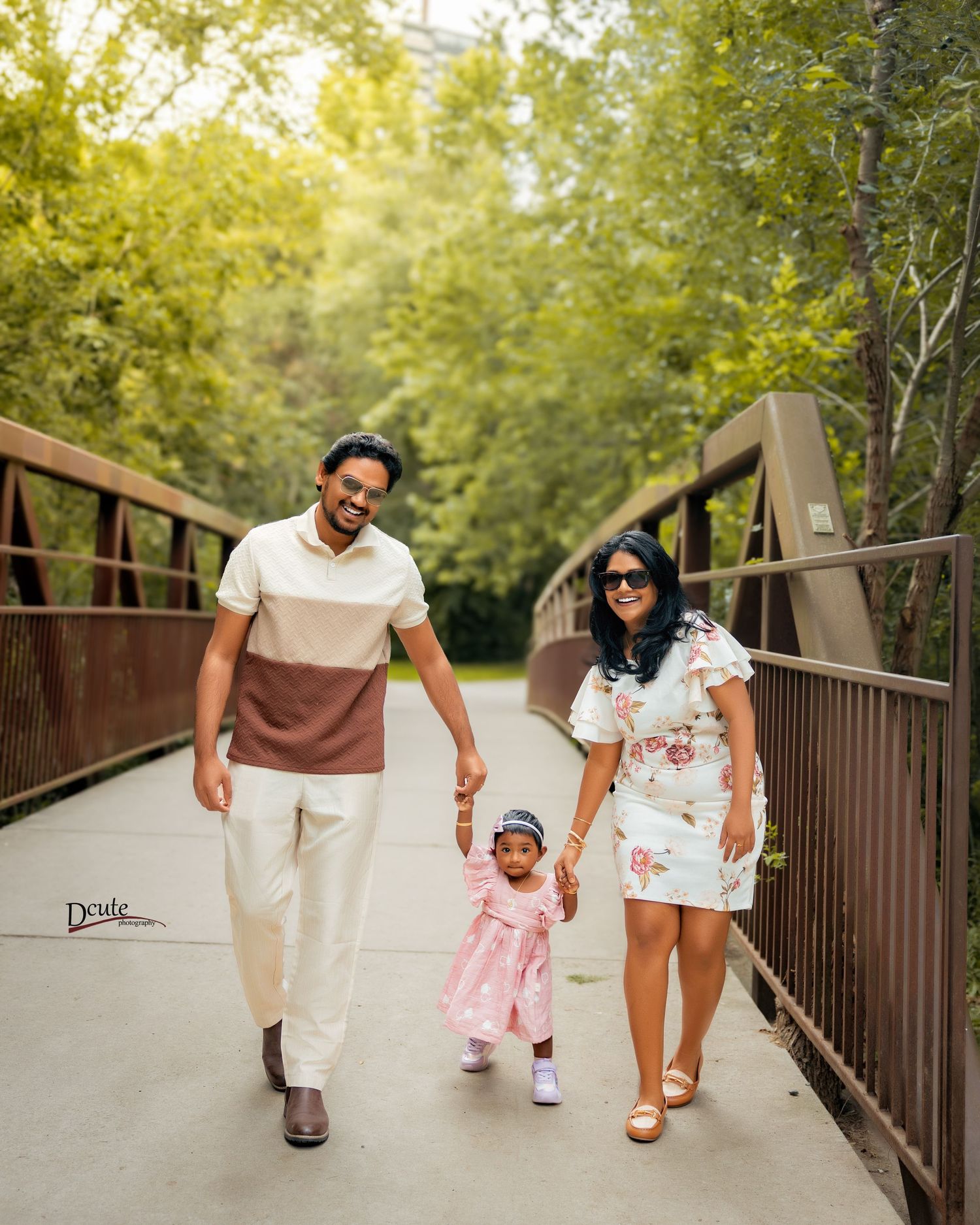 Happy South Asian family walking on wooden bridge, parents holding toddler girl in pink dress captured by Downtown Toronto family photograph