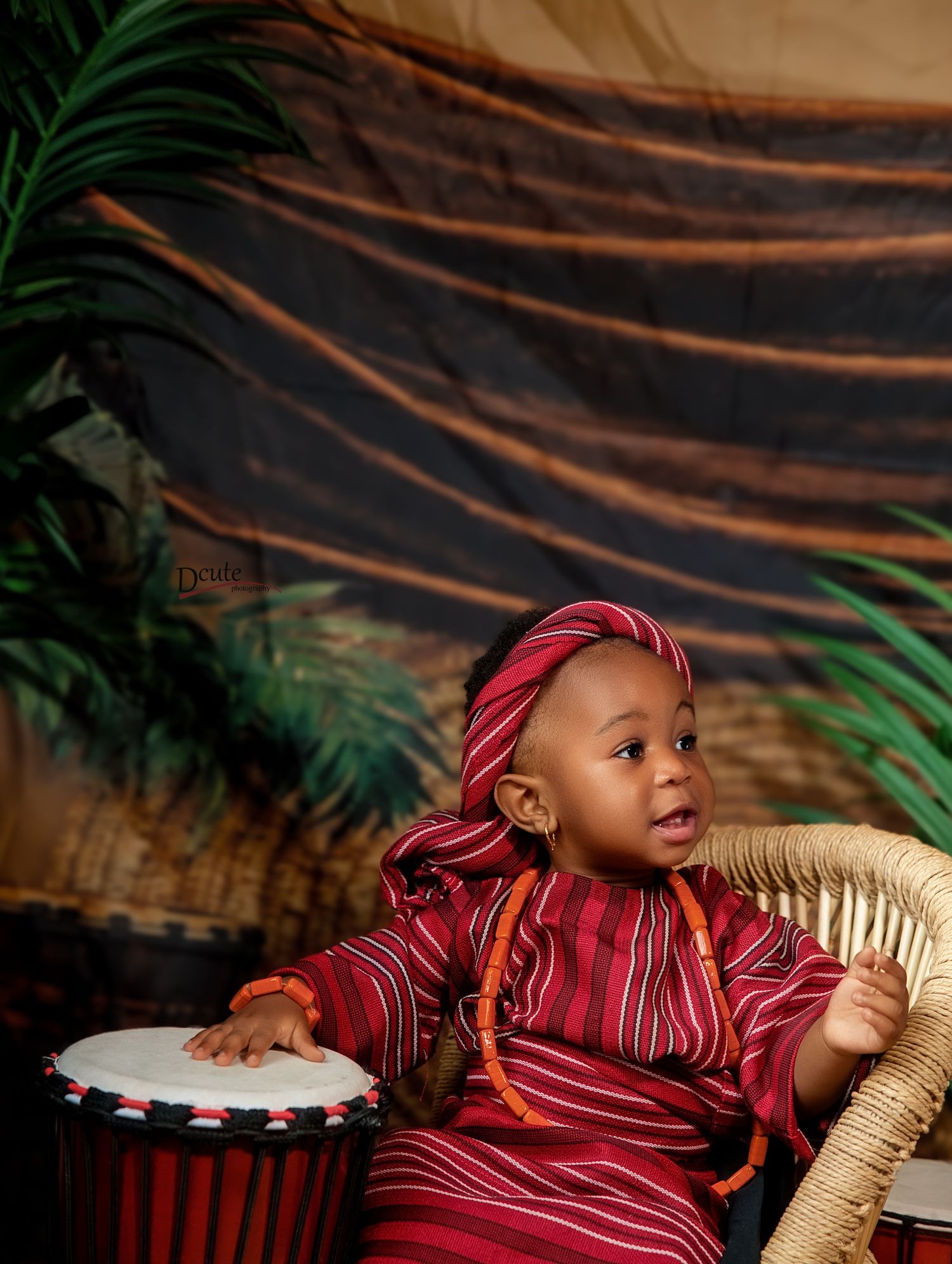 A toddler in red African attire plays a djembe drum while seated in a wicker chair with tropical backdrop.
