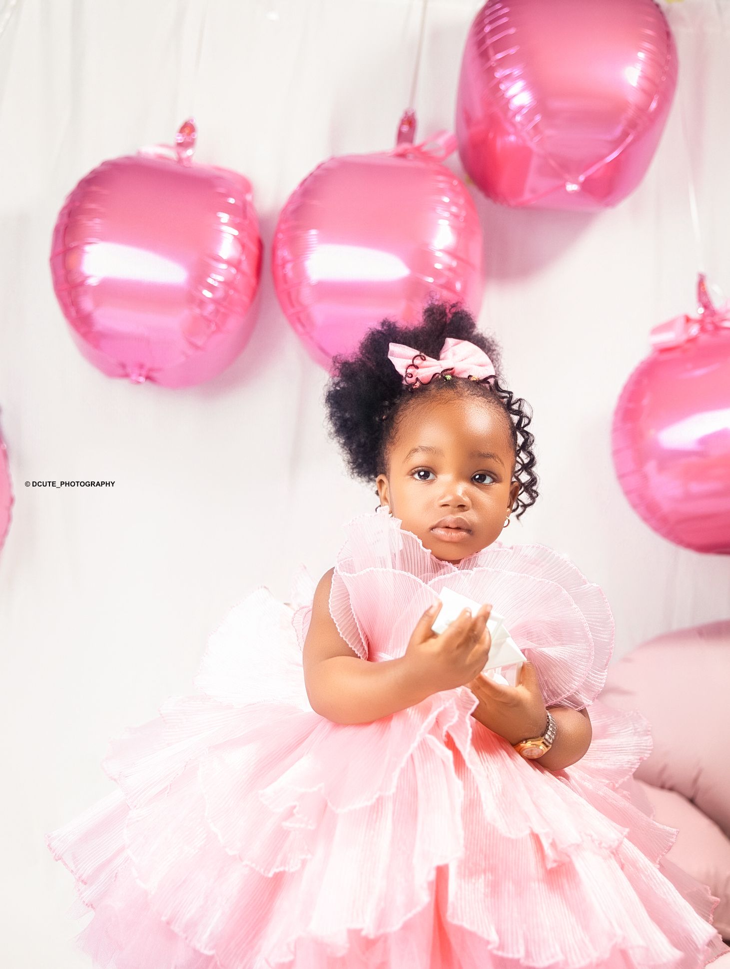 A little girl in a pink tutu dress stands near pink heart-shaped balloons at a birthday celebration.