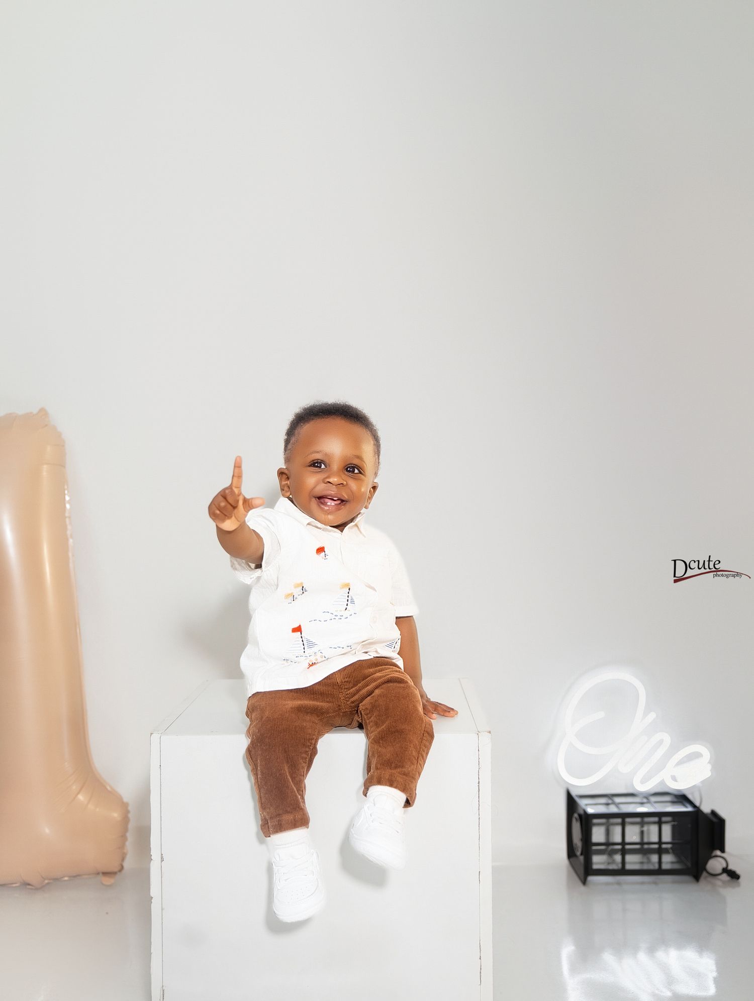 Smiling baby boy sitting on white box, holding up one finger, celebrating his 1st birthday with neon decor.