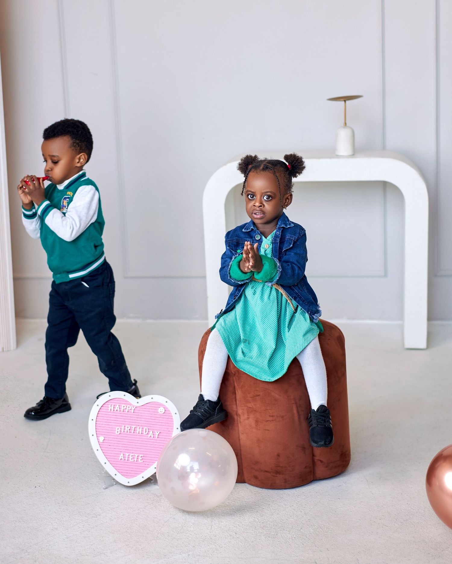 Two young children at a birthday party, girl sitting on ottoman in blue jacket and teal dress.