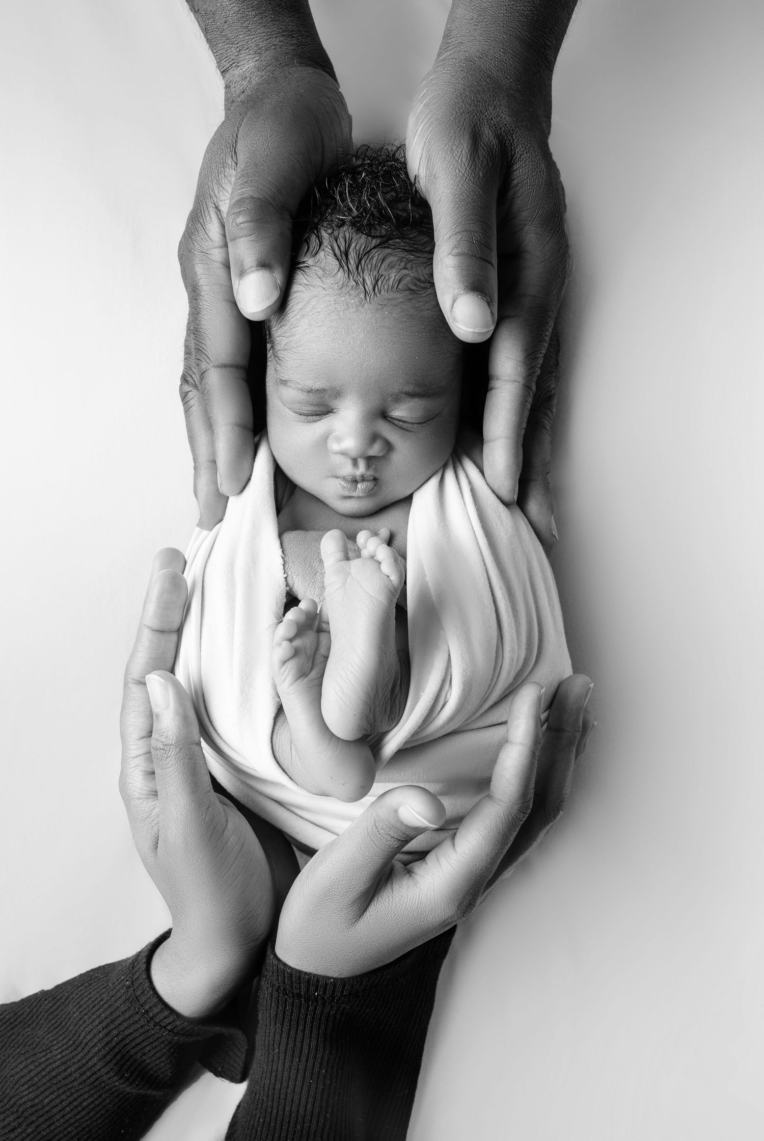 Black and white photo of a sleeping newborn baby cradled gently in two adult hands against a white background.