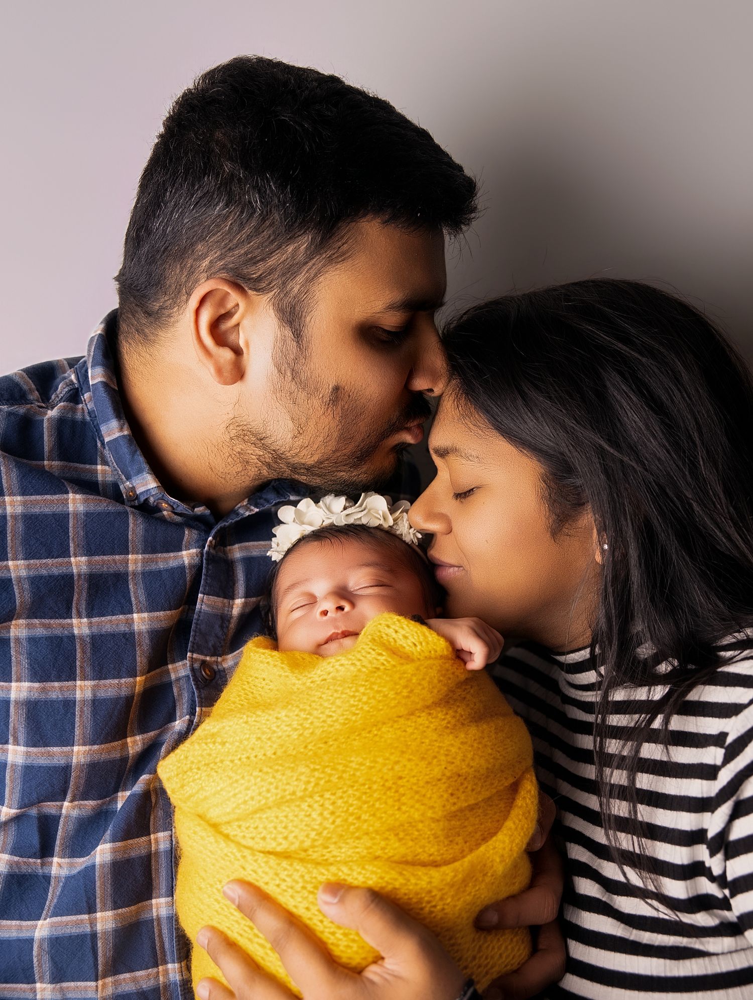 Parents kissing their sleeping newborn baby wrapped in a yellow swaddle blanket with a floral headband.