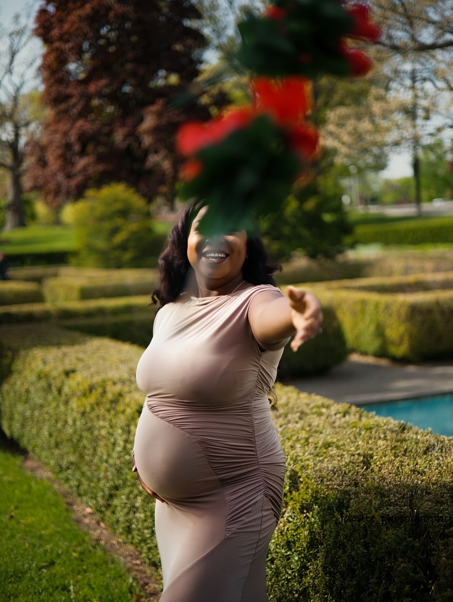 Pregnant woman in beige dress tossing red flowers in a sunny garden in a maternity photo session toronto