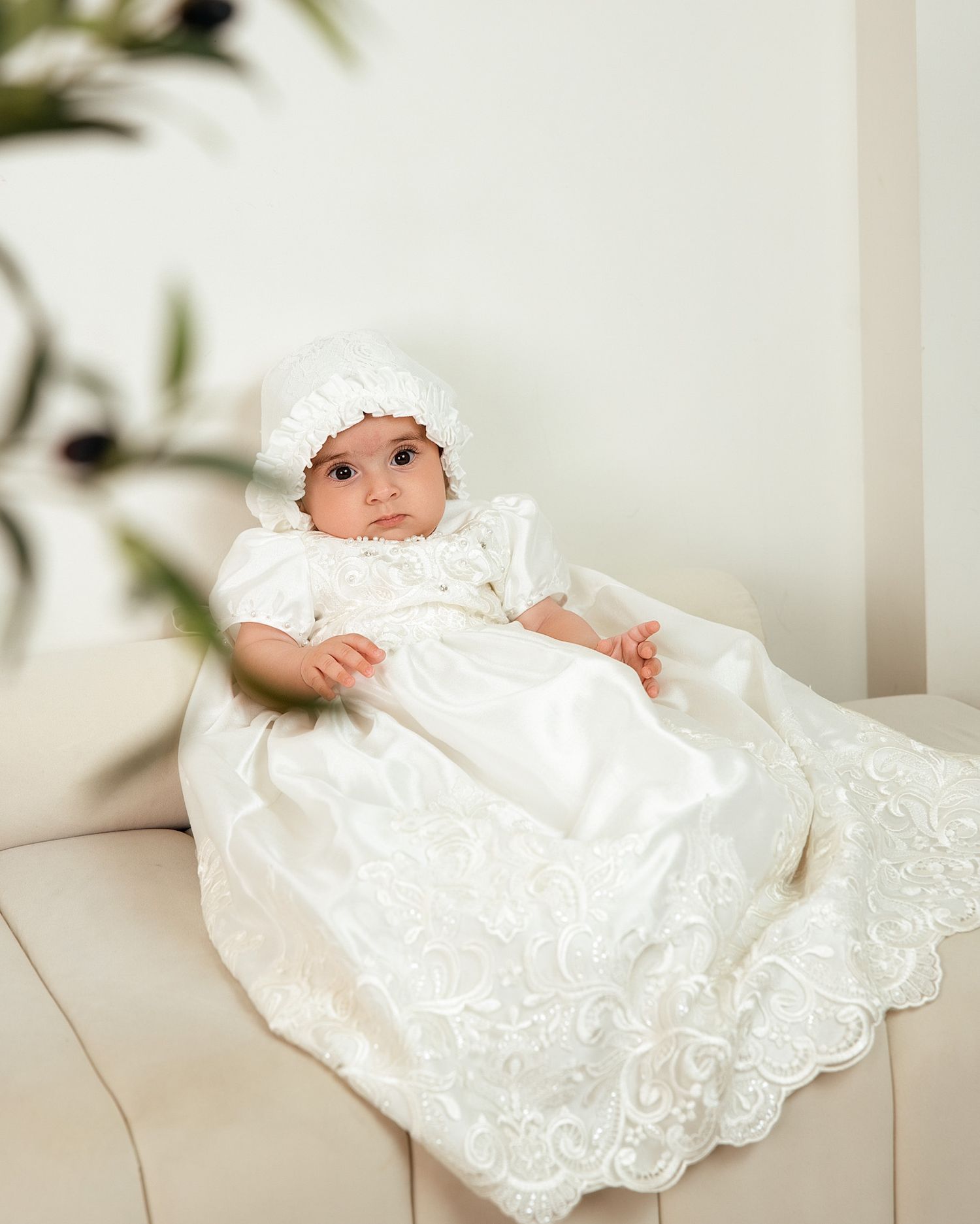A baby in an elegant white lace christening gown and bonnet sits on a cream sofa.