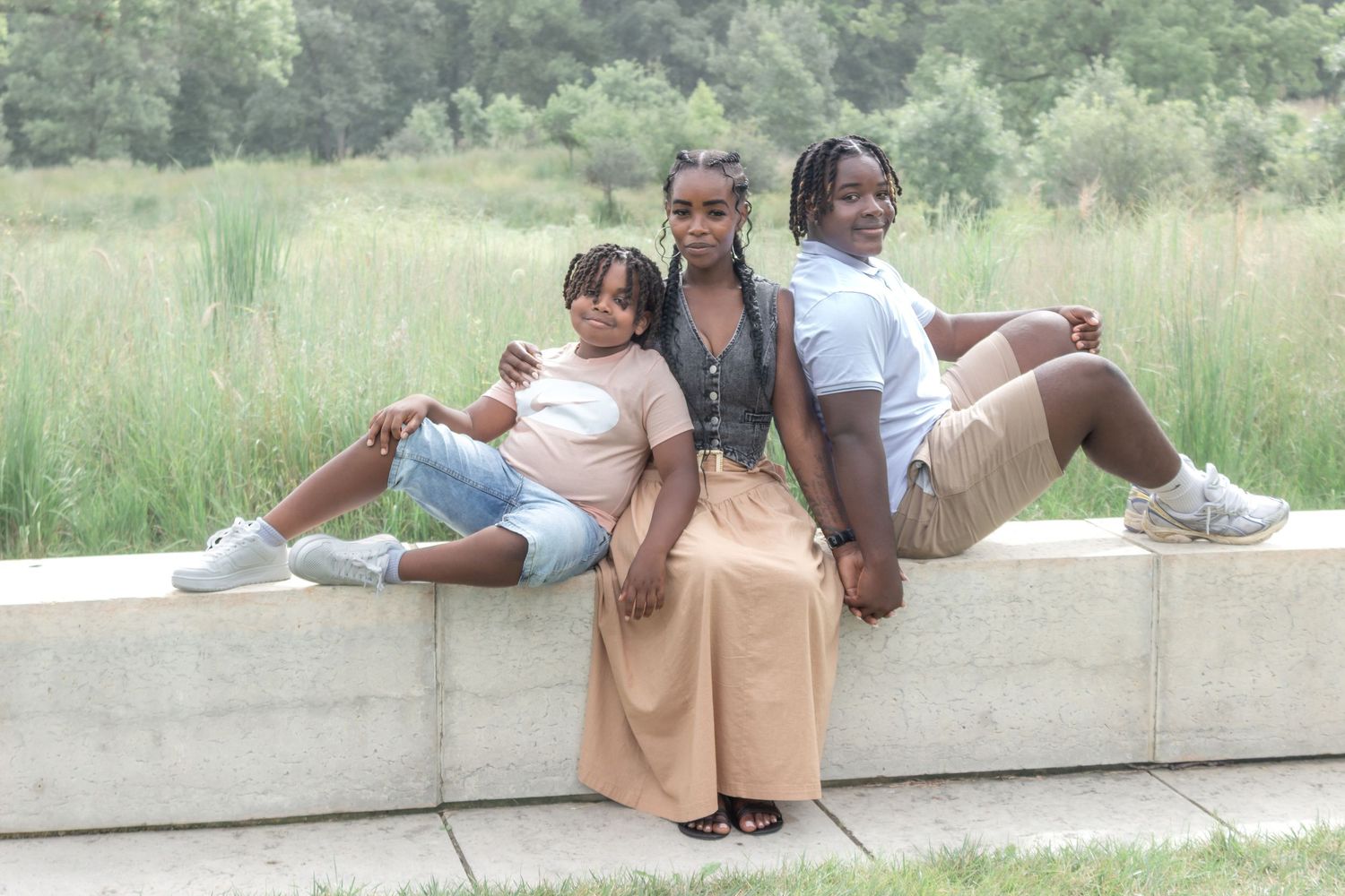 Mother sitting between her two children on a stone wall with a grassy field behind them during an outdoor family portrait session.