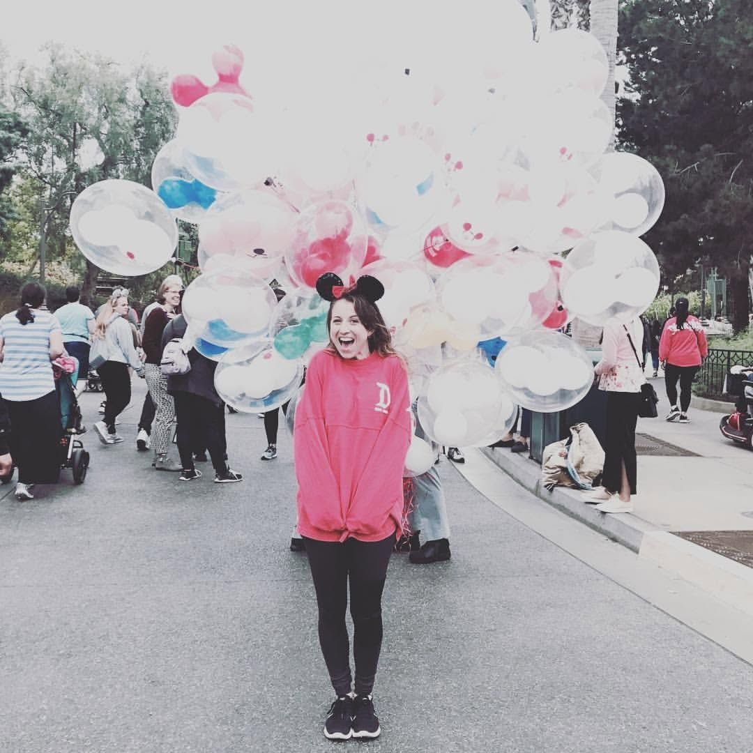 Woman in a pink dress standing in front of a colorful balloon-filled parade float during an outdoor celebration.