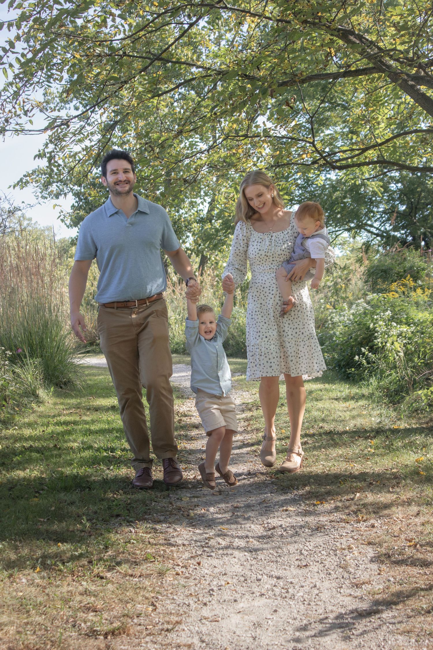 Parents walking hand in hand with their two young children along a sunlit wooded trail during an outdoor family photography session.