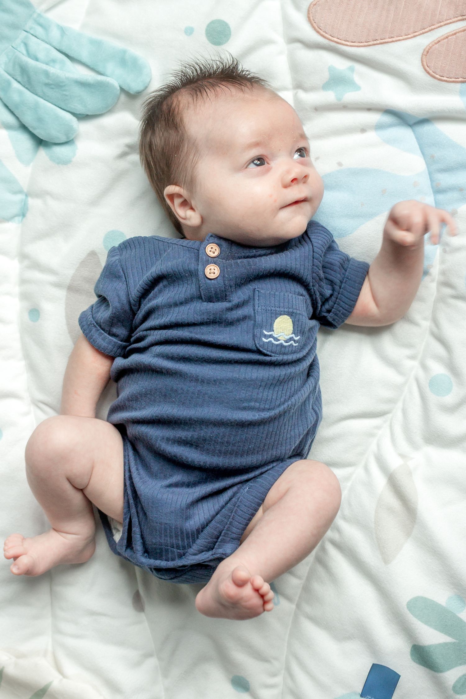 Baby boy in a blue onesie lying on a soft patterned blanket during a natural lifestyle baby photography session.