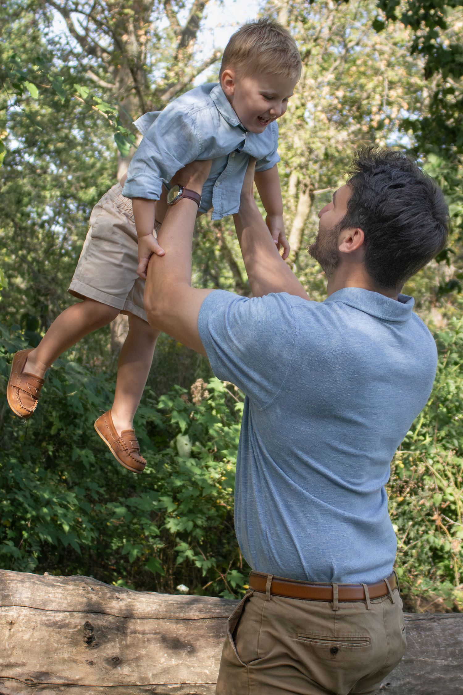 Father joyfully lifting his young son into the air beneath leafy trees during a playful outdoor family photography session.
