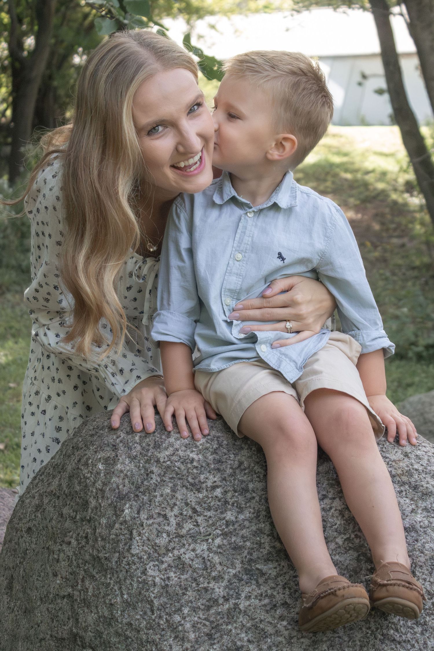 Young son kissing his smiling mother on the cheek while sitting together on a rock during an outdoor family photography session.