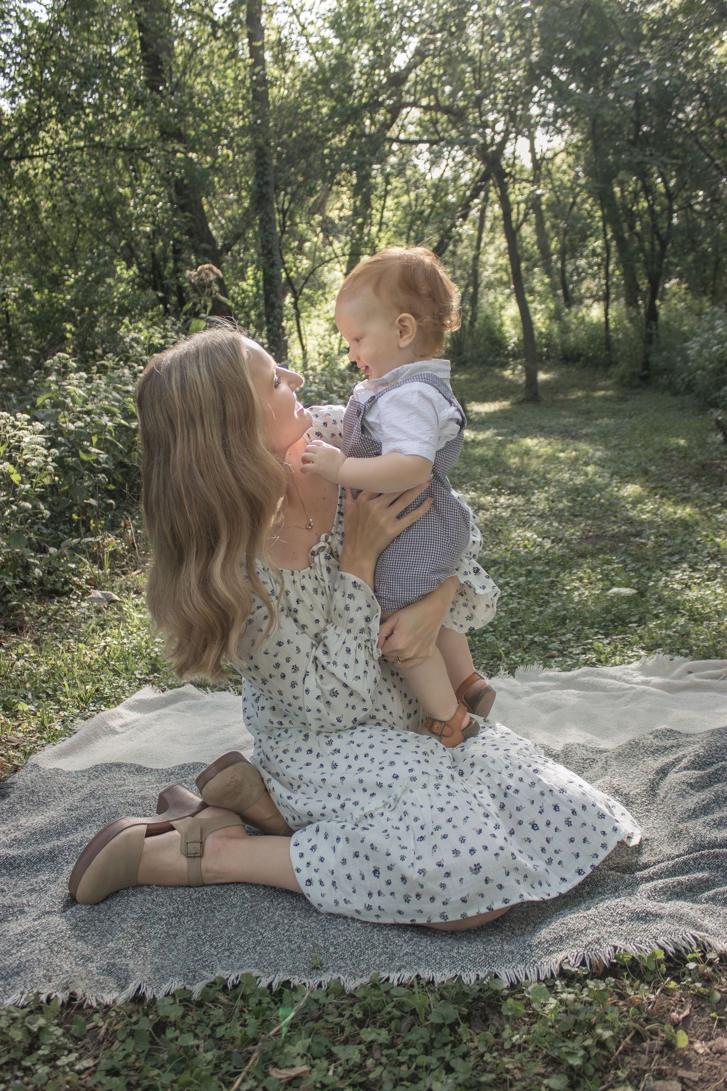 Mother in a floral dress holding her young child on a blanket in a wooded park during a tender outdoor family photography session.