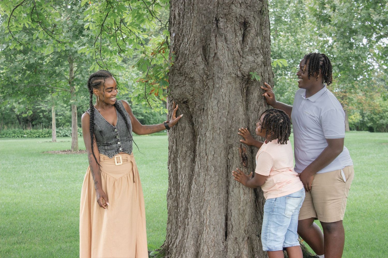 Mother smiling as her two children play peekaboo around a large tree during a candid outdoor family photography session.