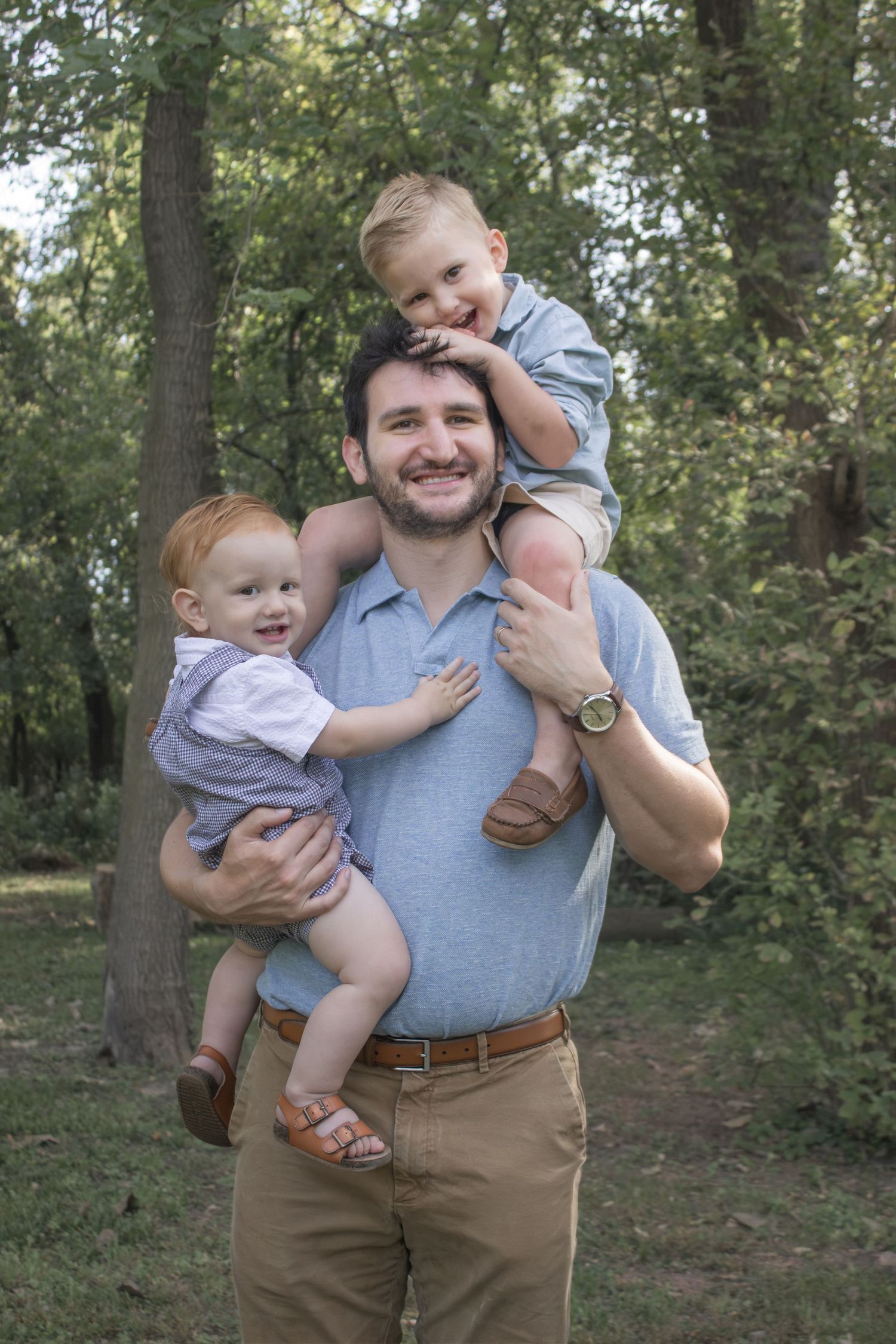 Smiling father carrying one child on his shoulders and another in his arms during a playful outdoor family portrait in a wooded park.