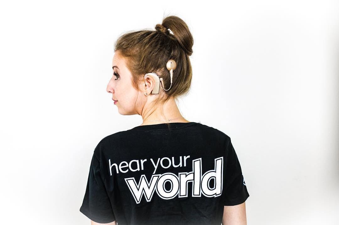 Woman with hair in a bun wearing a black Hear Your World T-shirt, photographed from behind against a white background.
